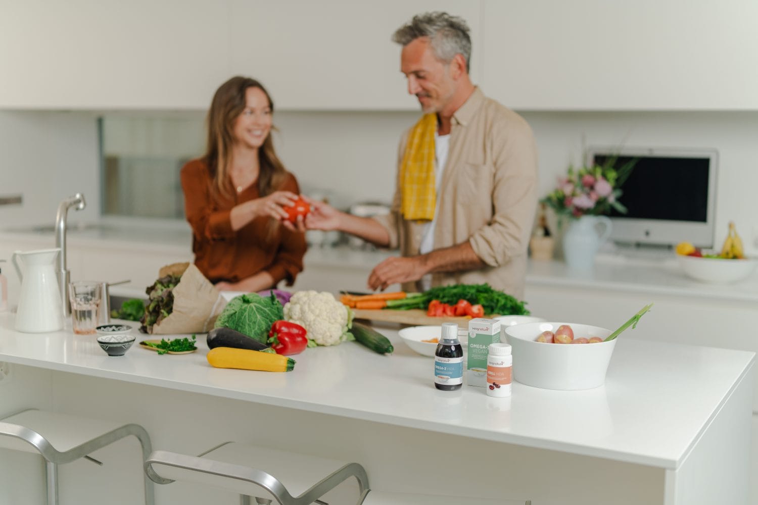 Ein Mann und eine Frau lächeln und bereiten gemeinsam Gemüse in einer hellen Küche zu. Frische Produkte, Nahrungsergänzungsmittel und eine Schale Äpfel stehen auf der Theke im Vordergrund und schaffen eine lebendige Szene.