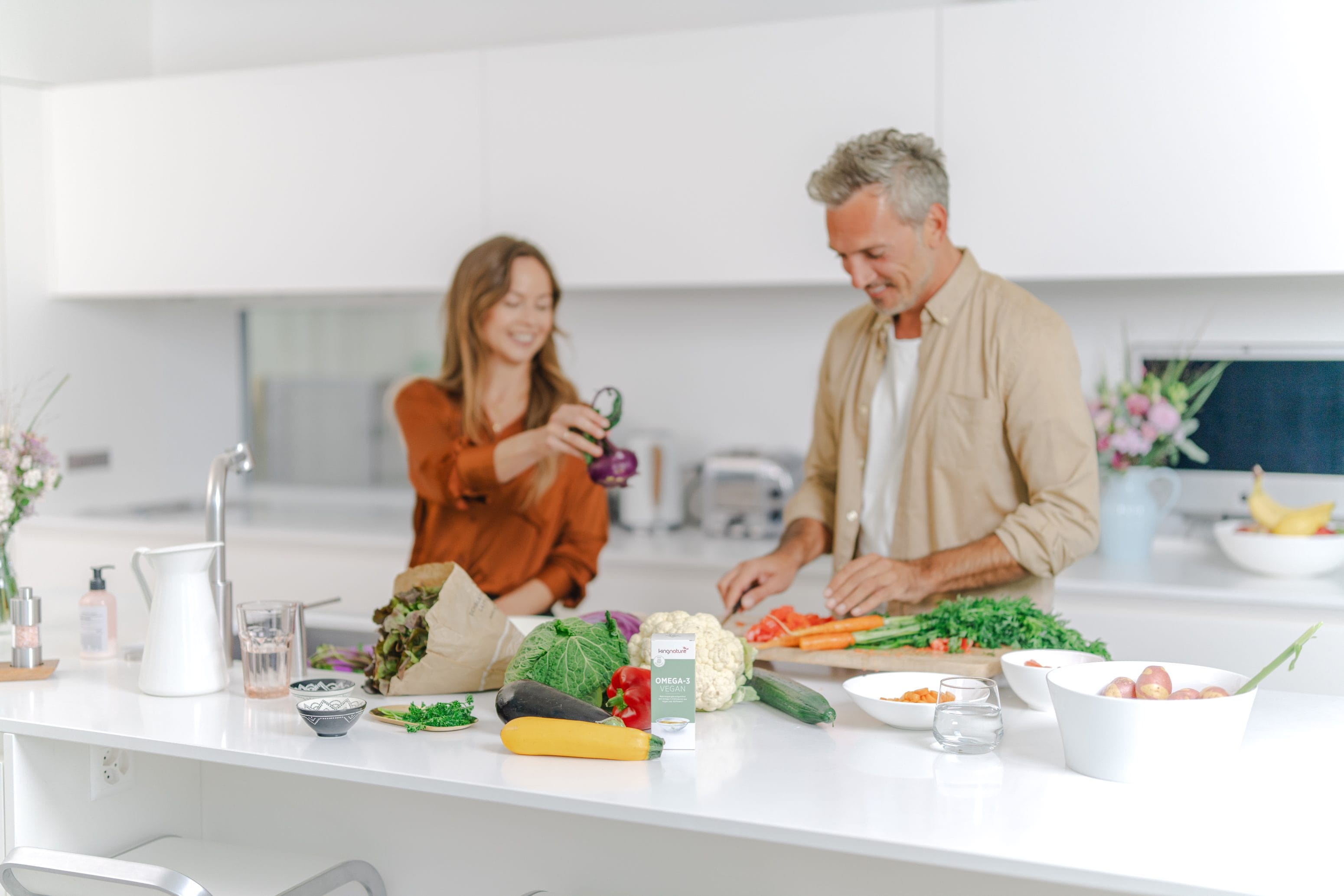 Ein Mann und eine Frau lächeln, während sie gemeinsam in einer hellen, modernen Küche Gemüse zubereiten. Frische Produkte und Bio-Zutaten liegen auf der Küchentheke ausgebreitet.