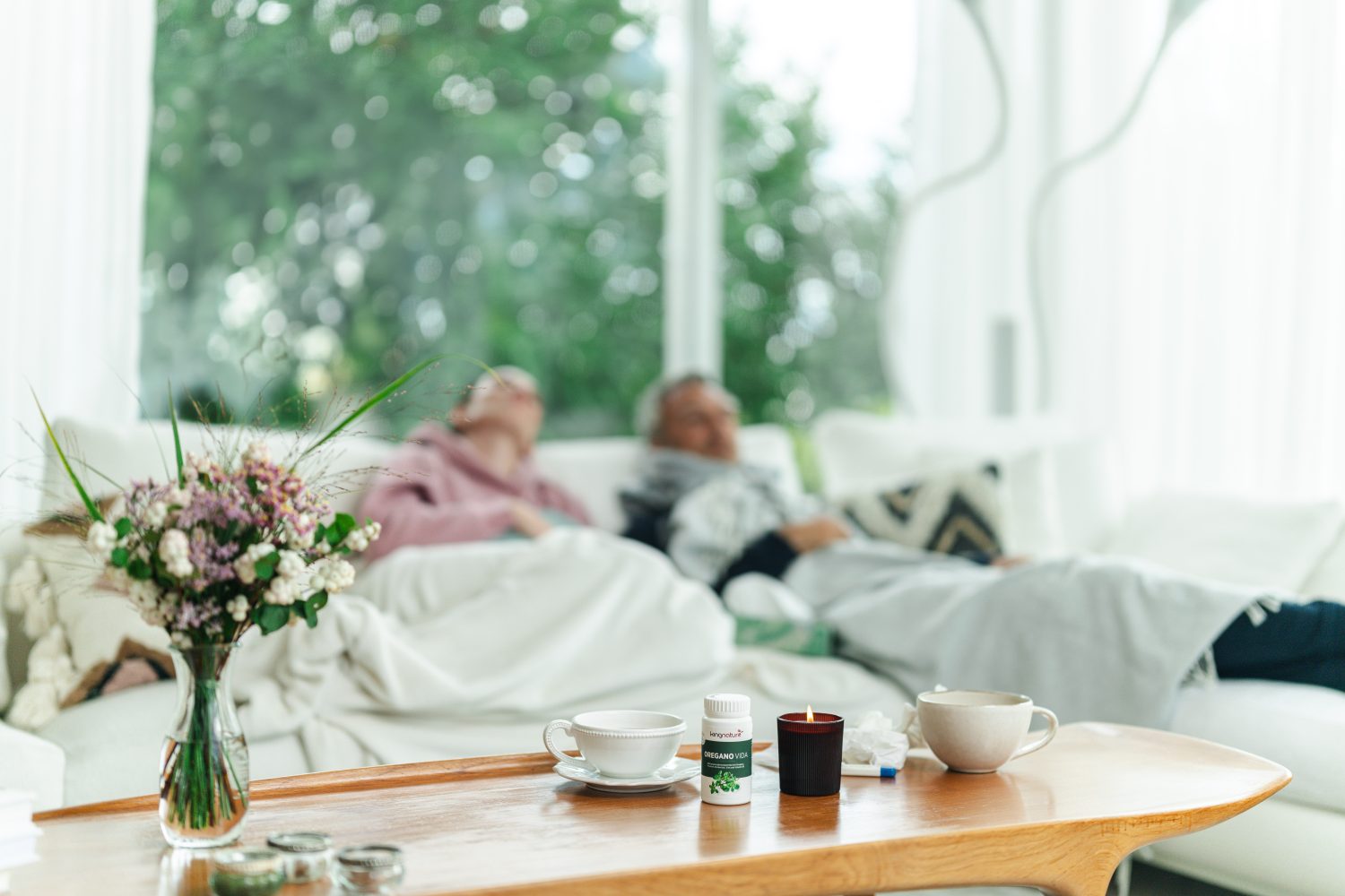 Eine Nahaufnahme eines hölzernen Couchtischs mit einer Tasse, einer Oregano-Vida-Kaufflasche, einer Kerze und Blumen. Im unscharfen Hintergrund ruhen zwei Personen unter Decken auf einem Sofa neben großen, hellen Fenstern.