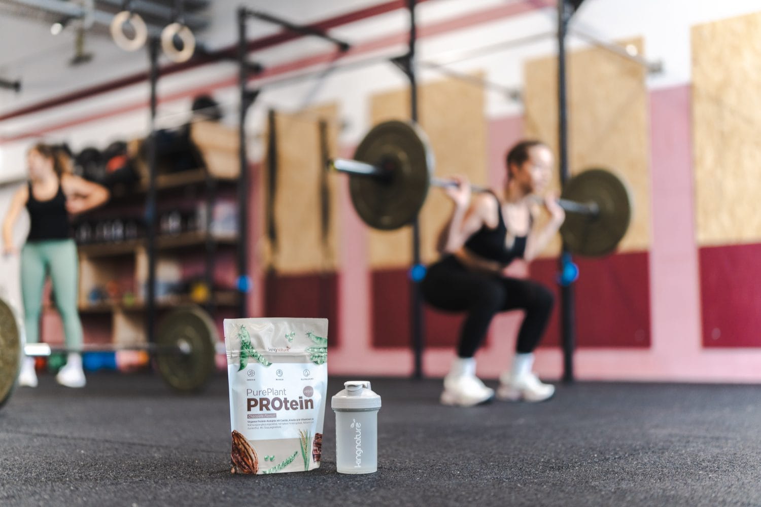 A container of protein powder and a shaker bottle are lying on the gym floor, while in the background, a person surrounded by exercise equipment and weights is doing barbell squats.
