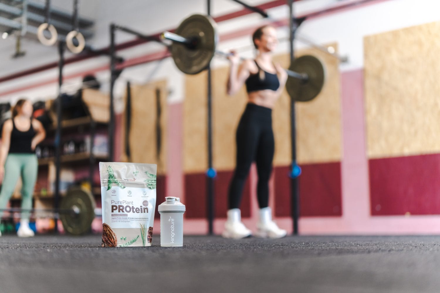 Eine Tüte Proteinpulver und ein Shaker stehen auf dem Boden des Fitnessstudios, während im unscharfen Hintergrund eine Frau eine Hantel stemmt. In der Nähe steht eine weitere Person neben einem Fitnessgerät.