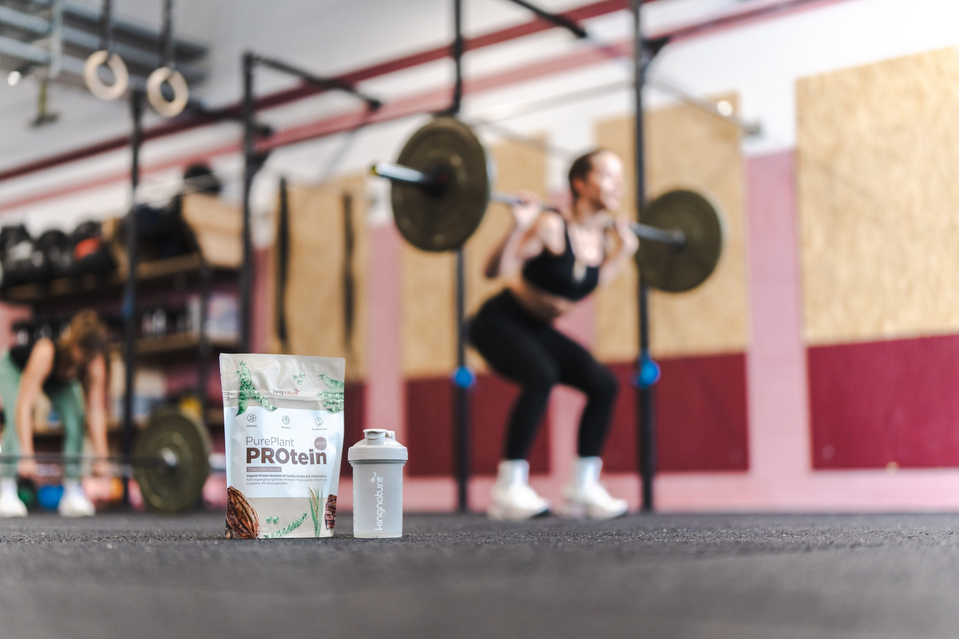A bag of protein powder and a shaker bottle are lying on the floor of a gym. In the background, a blurred figure can be seen lifting a barbell next to squat racks. Buy Amino Vida online now and support your fitness routine.