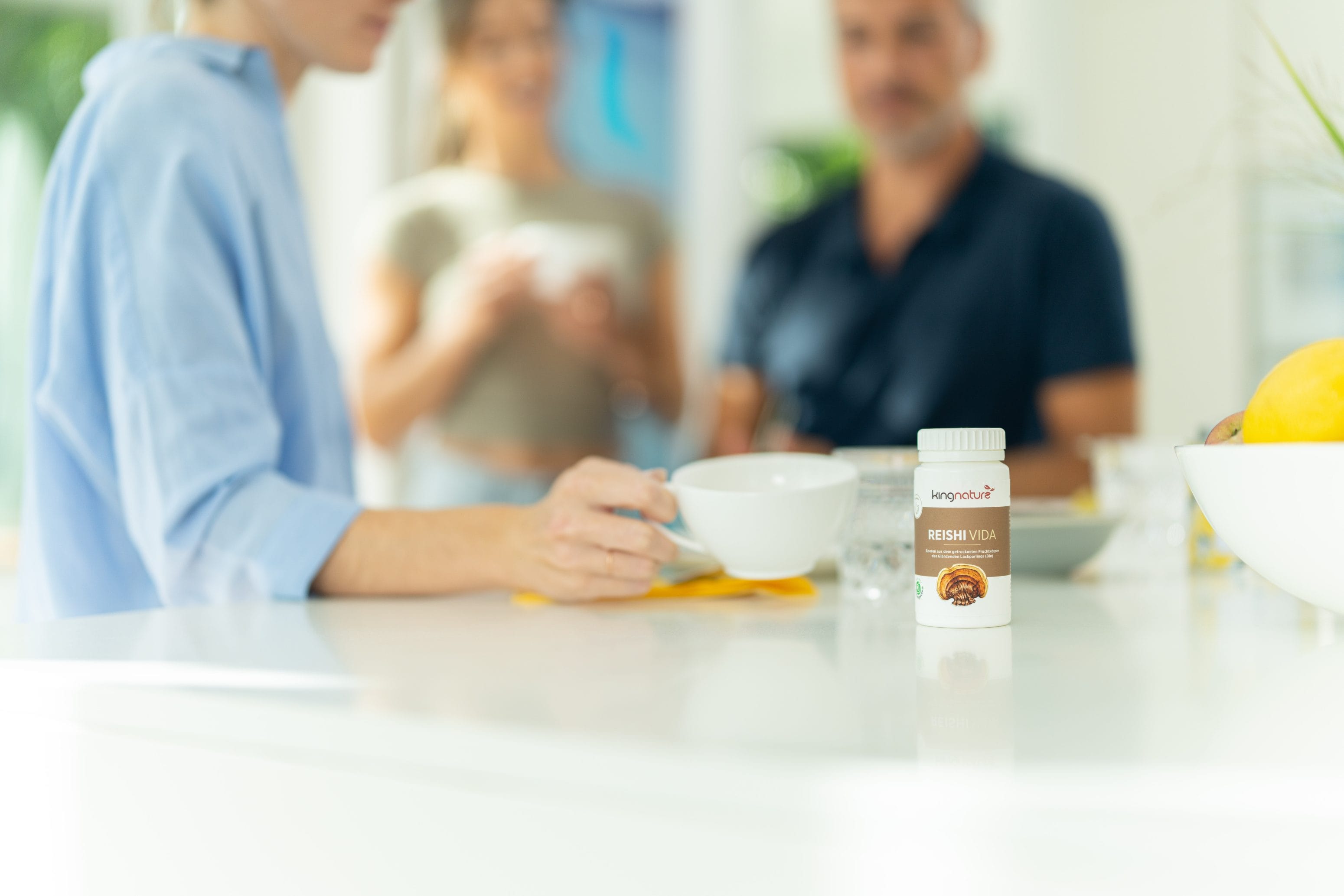 Eine Person mit einer weißen Tasse sitzt an einer hellen Küchentheke. Im Fokus steht eine Flasche Reishi Vita-Nahrungsergänzungsmittel. Zwei weitere Personen unterhalten sich im unscharfen Hintergrund und unterstreichen die freundliche Atmosphäre.