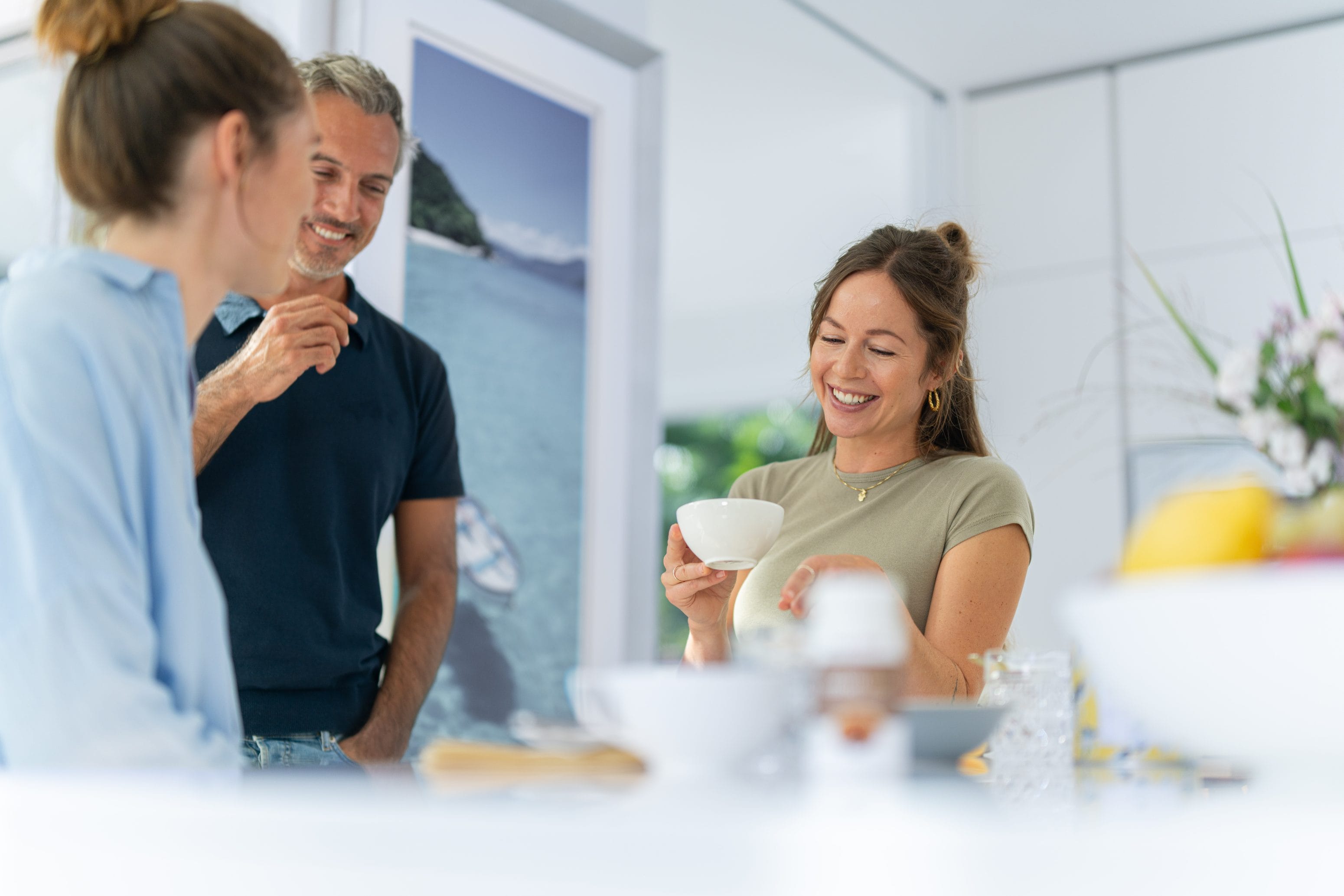 Drei Menschen stehen lächelnd und unterhalten sich in einer hellen Küche. Eine Frau hält eine weiße Tasse in der Hand und lacht, während die anderen beiden zuhören und das lockere und gesellige Beisammensein genießen.
