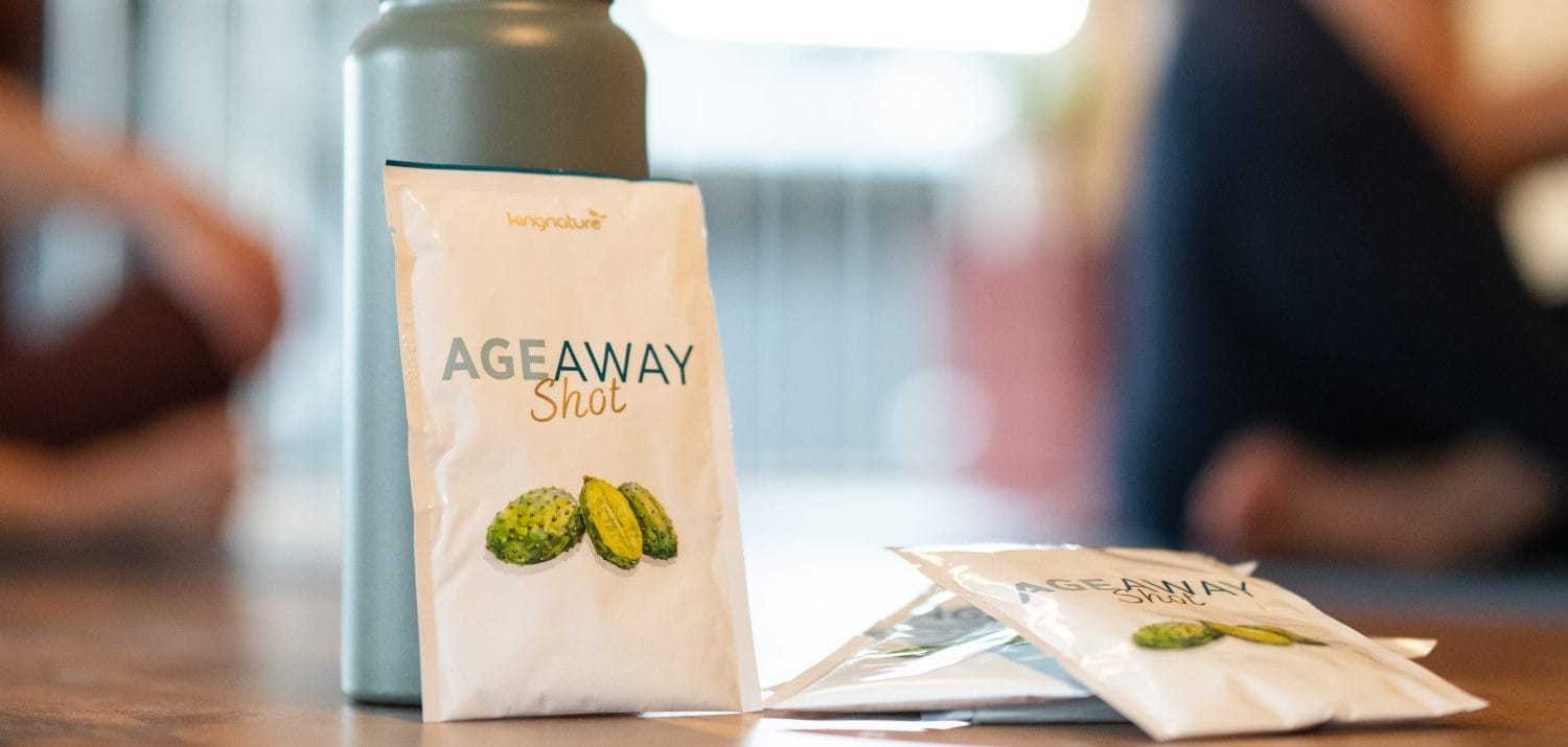 Eine Nahaufnahme von AgeAway Shot-Päckchen mit grünen Fruchtillustrationen, die neben einer grauen Wasserflasche auf einer Holzoberfläche platziert sind; unscharfer Hintergrund mit sitzenden Personen.