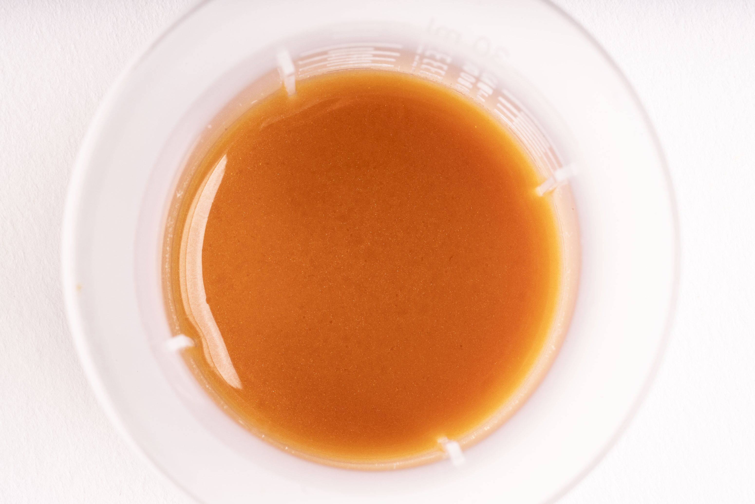 Draufsicht auf einen klaren Messbecher mit einer orangefarbenen Flüssigkeit vor einem weißen Hintergrund. Die Flüssigkeit füllt etwa ein Drittel des Bechers aus, und an der Seite sind Messlinien zu sehen.