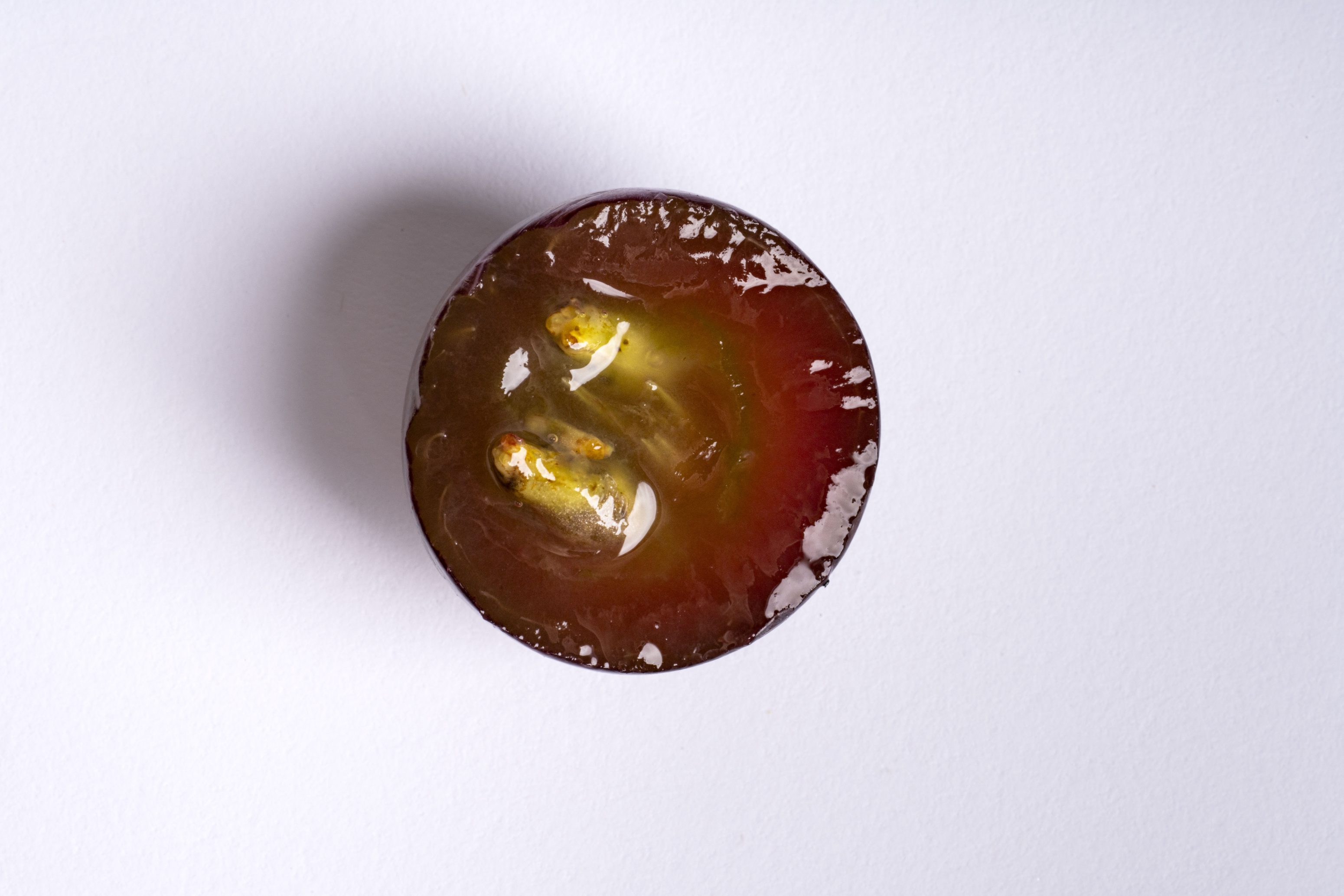 Nahaufnahme einer aufgeschnittenen roten Weintraube von oben, mit durchscheinendem Fruchtfleisch und zwei Kernen im Inneren, vor einem schlichten weißen Hintergrund.