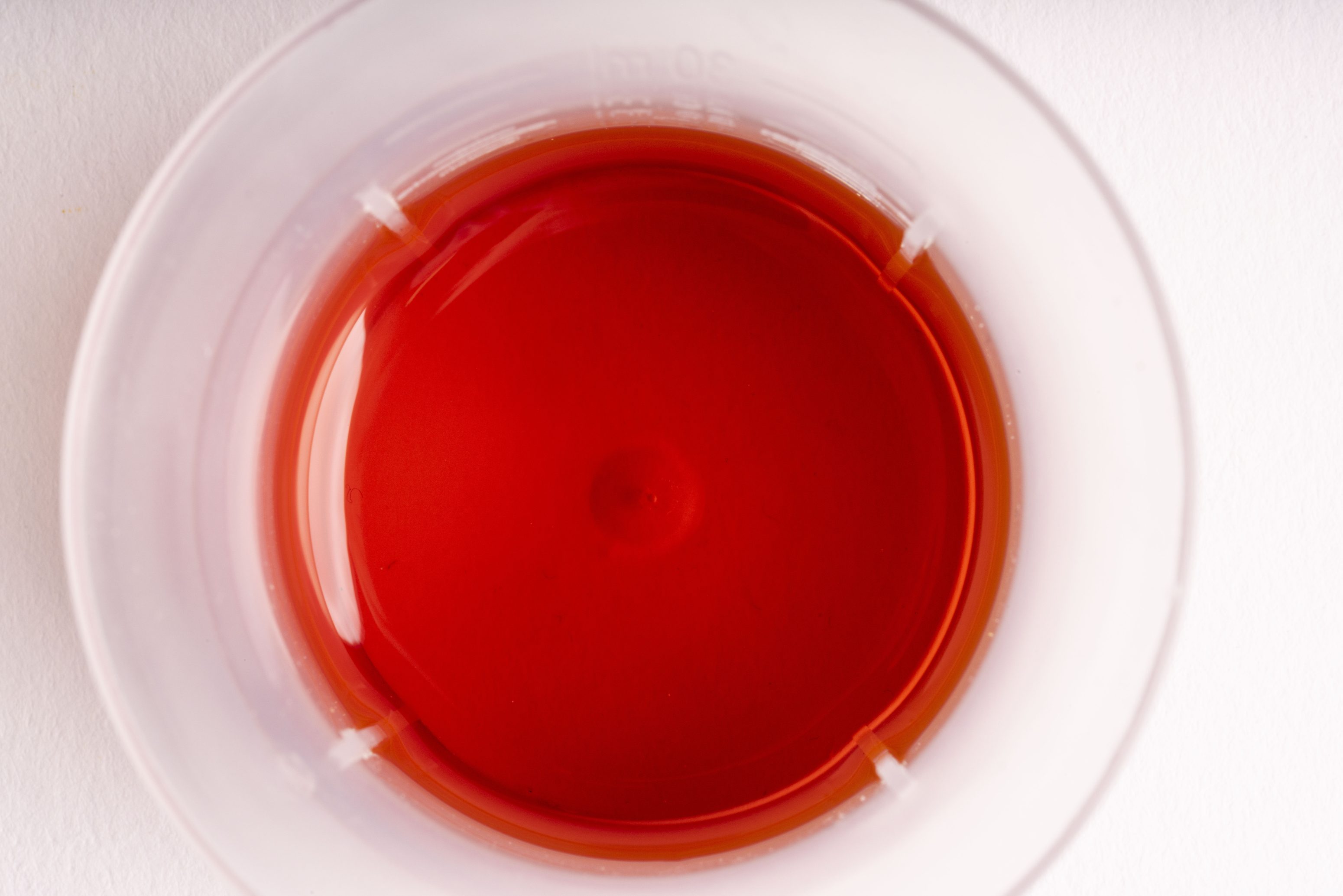 Draufsicht auf einen klaren Plastikbecher, der mit einer leuchtend roten Flüssigkeit gefüllt ist und auf einer weißen Fläche steht.