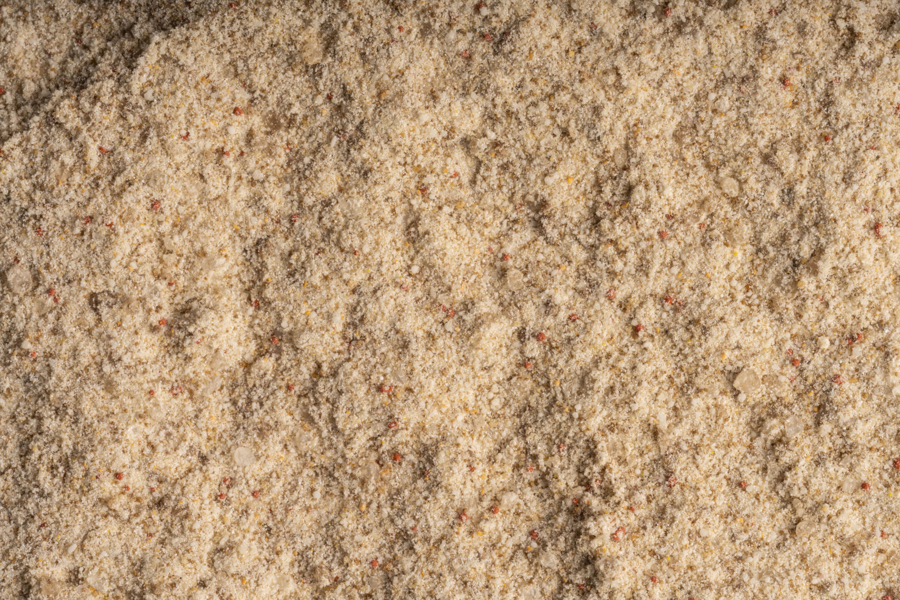 Nahaufnahme einer strukturierten, hellbraunen, pulverförmigen Substanz mit kleinen, verstreuten roten und gelben Flecken.