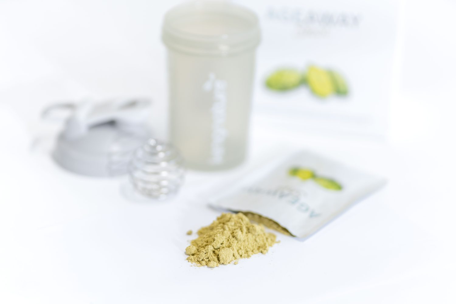 Ein Haufen hellbraunes Pulver liegt auf einer weißen Fläche neben einer geöffneten Packung. Im Hintergrund befinden sich eine Shakerflasche, eine Metallmischkugel und eine Schachtel mit grünen Früchten.