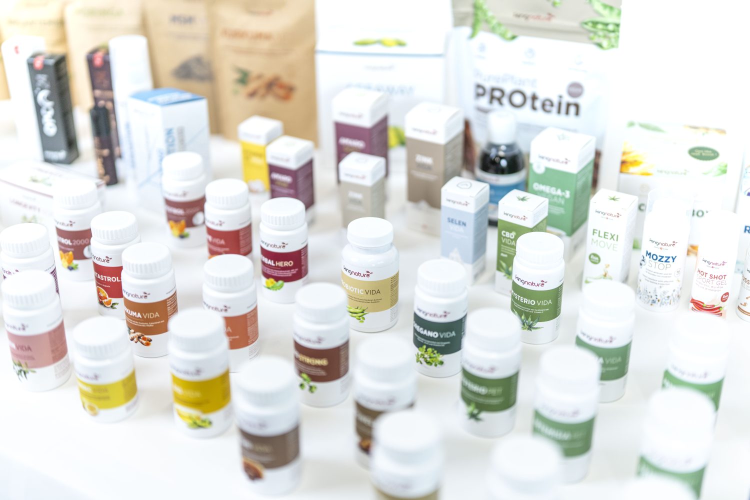 Mehrere weiße Nahrungsergänzungsflaschen und -schachteln mit bunten Etiketten sind auf einer weißen Fläche angeordnet. Zu den Produkten gehören pflanzliche Proteine, Vitamine und andere Nahrungsergänzungsmittel. Der Hintergrund ist hell und unscharf.