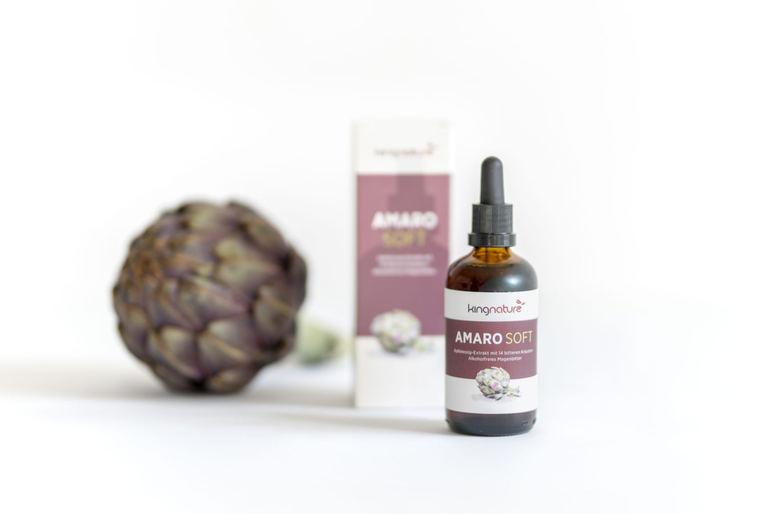 Eine Tropfflasche aus braunem Glas mit der Aufschrift "Amaro Soft" steht vor einer passenden Schachtel, auf der linken Seite eine ganze Artischocke, alles vor einem weißen Hintergrund.