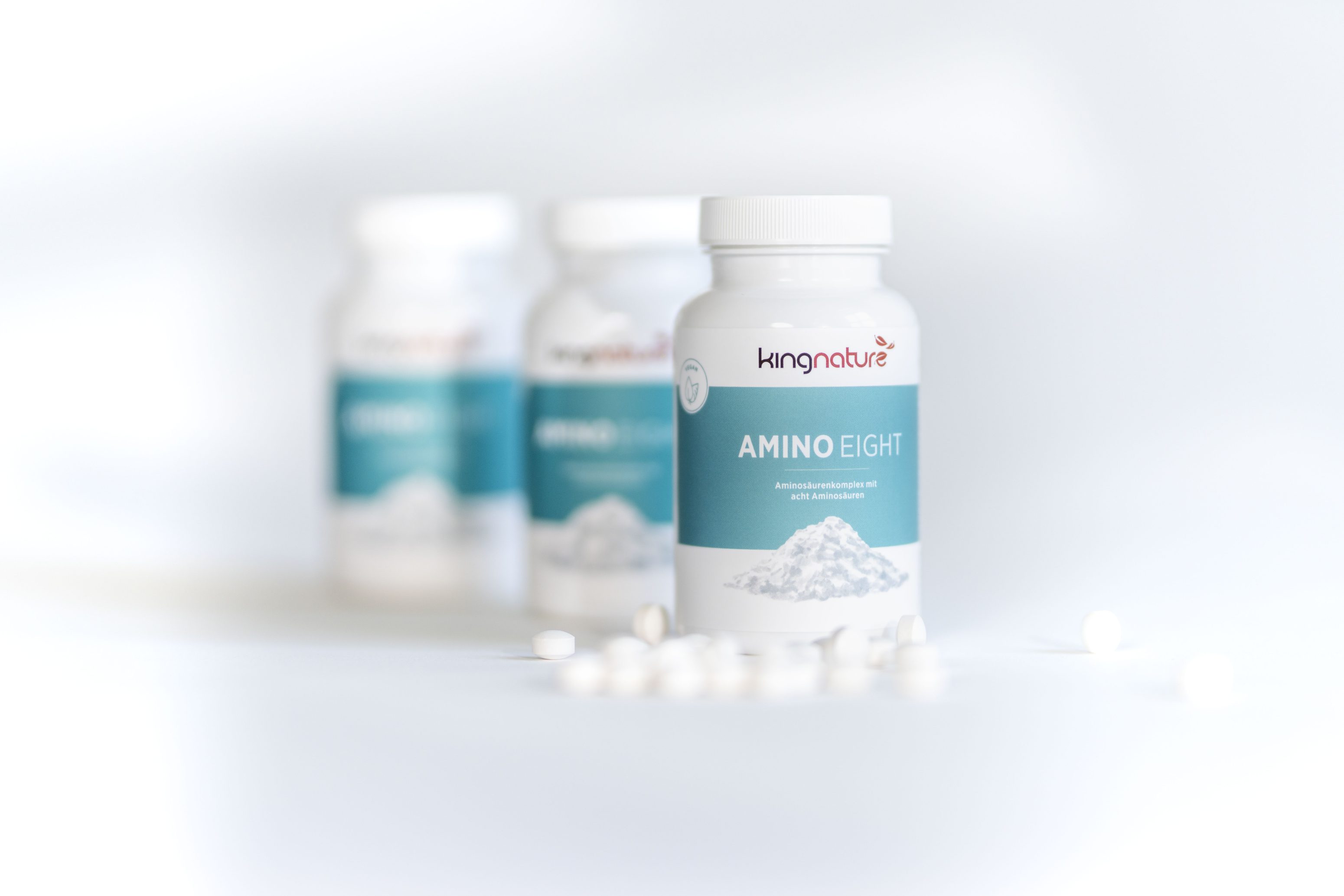 Drei weiße Flaschen mit Kingnature Amino Eight Nahrungsergänzungsmitteln sind auf einer weißen Fläche angeordnet, wobei eine Flasche im Vordergrund scharf und zwei im Hintergrund unscharf zu sehen sind. Mehrere weiße Tabletten liegen verstreut herum.