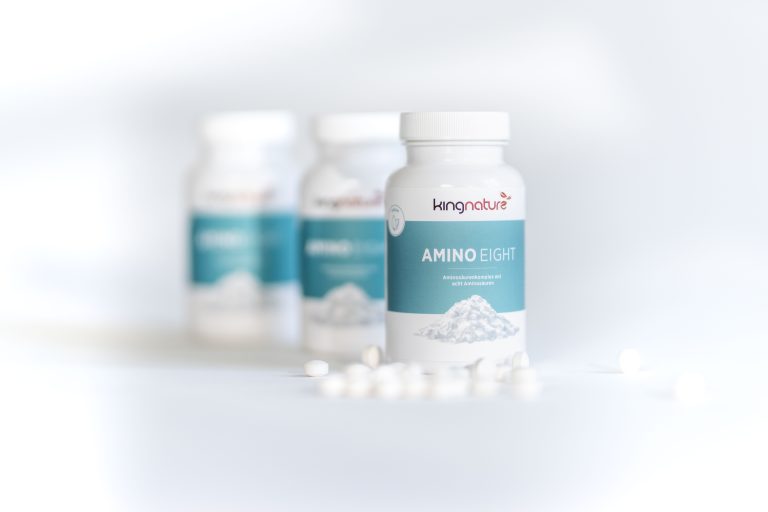 Drei weiße Flaschen mit Kingnature Amino Eight Nahrungsergänzungsmitteln sind auf einer weißen Fläche angeordnet, wobei eine Flasche im Vordergrund scharf und zwei im Hintergrund unscharf zu sehen sind. Mehrere weiße Tabletten liegen verstreut herum.