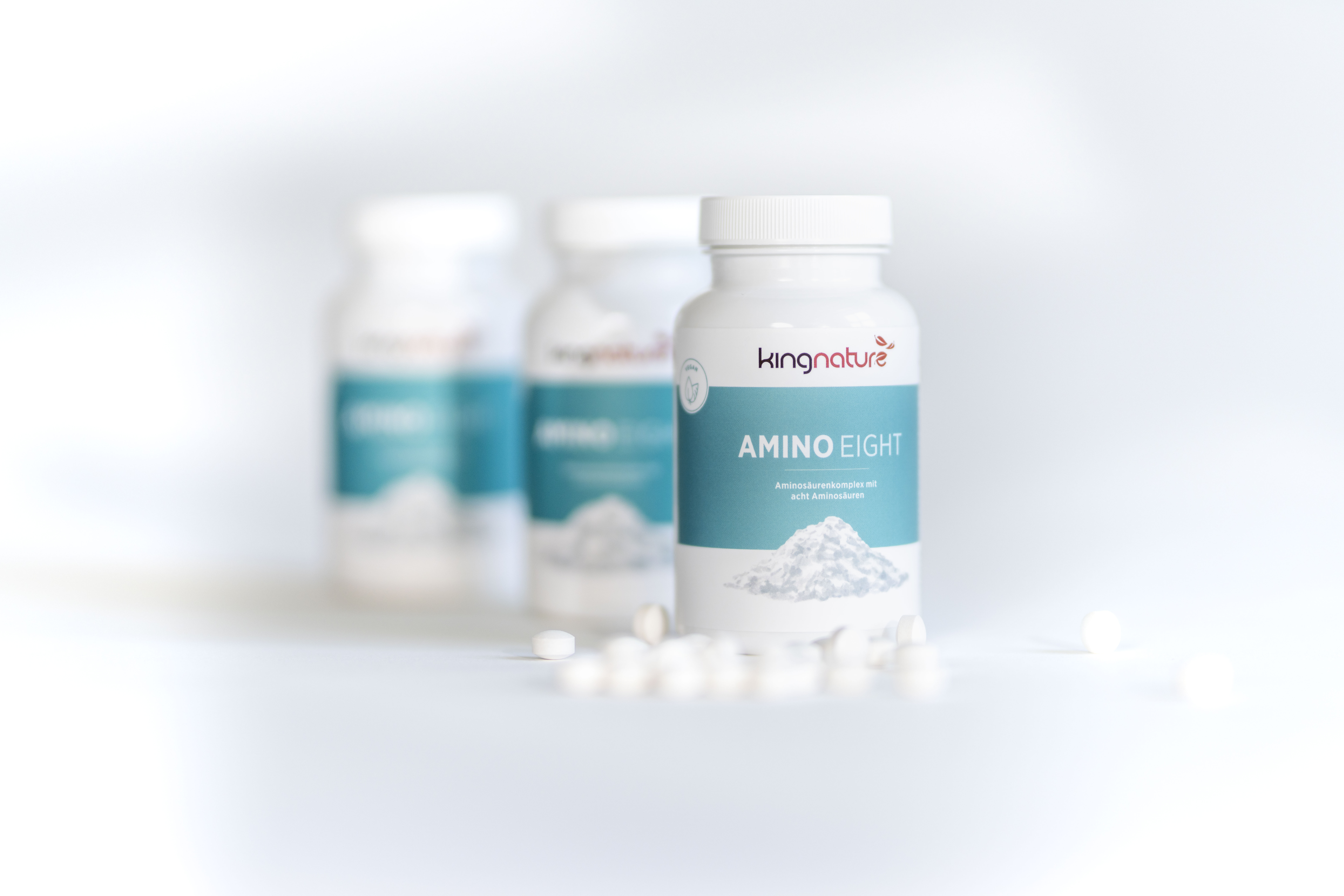 Drei weiße Flaschen mit Kingnature Amino Eight Nahrungsergänzungsmitteln sind auf einer weißen Fläche angeordnet, wobei eine Flasche im Vordergrund scharf und zwei im Hintergrund unscharf zu sehen sind. Mehrere weiße Tabletten liegen verstreut herum.