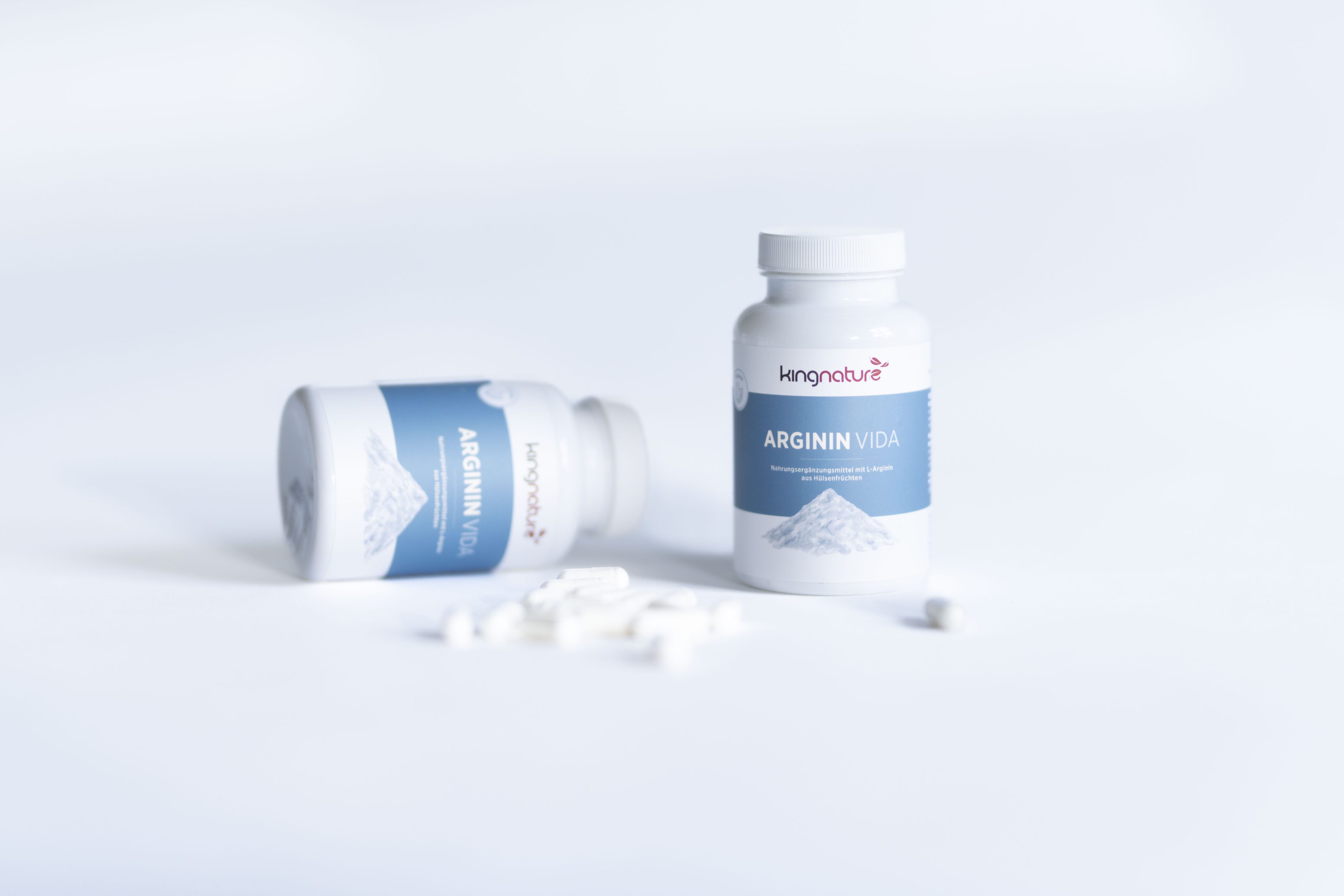 Zwei weiße Flaschen mit der Aufschrift kingnature ARGININI VIDA stehen auf einer weißen Fläche, eine aufrecht und eine liegend, mit mehreren weißen Kapseln, die in der Nähe verstreut sind. Der Hintergrund ist minimal und hell.
