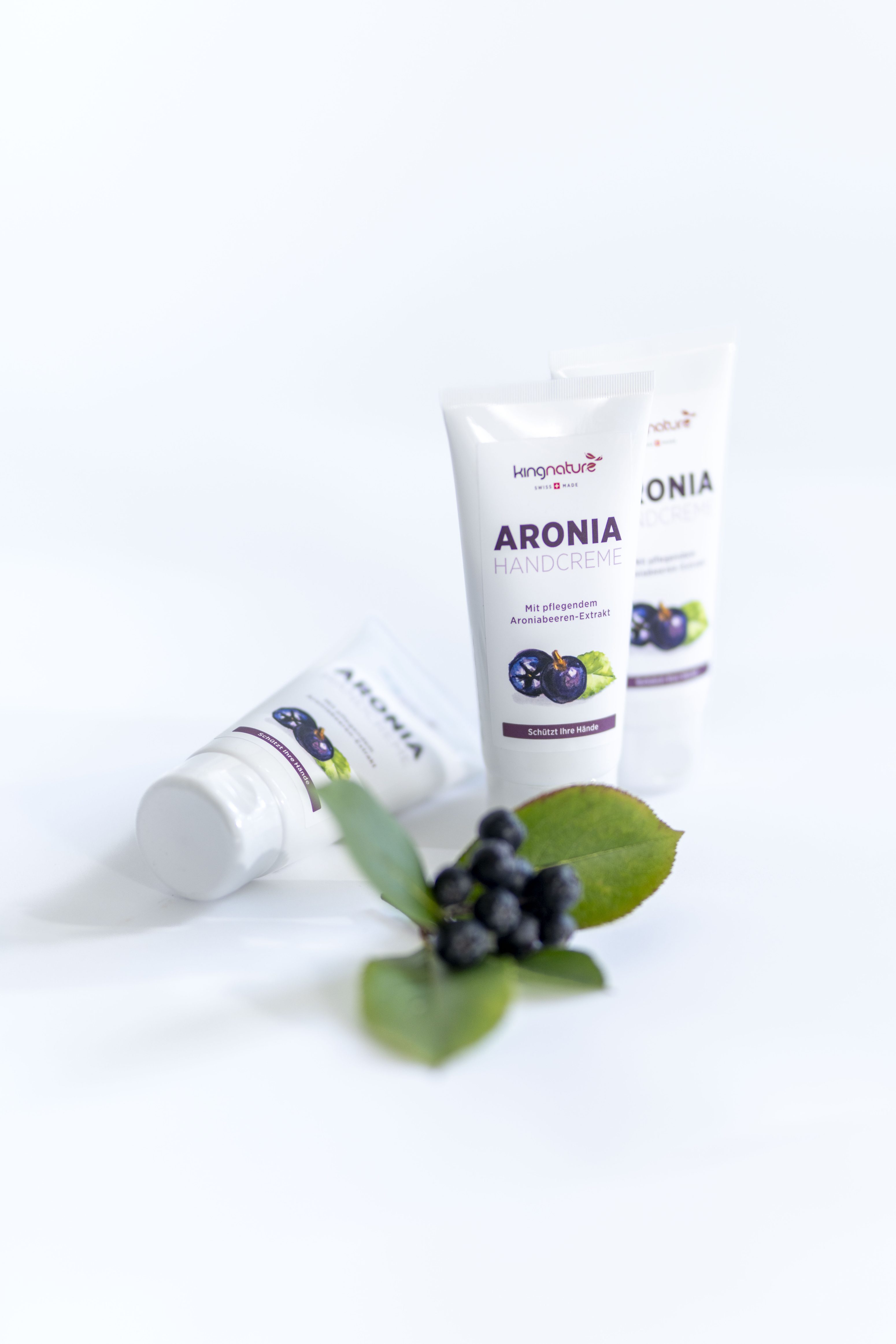 Drei Tuben Aronia-Handcreme sind auf einer weißen Fläche angeordnet, wobei zwei aufrecht stehen und eine liegt. Davor befinden sich ein Zweig mit frischen Aroniabeeren und grüne Blätter.