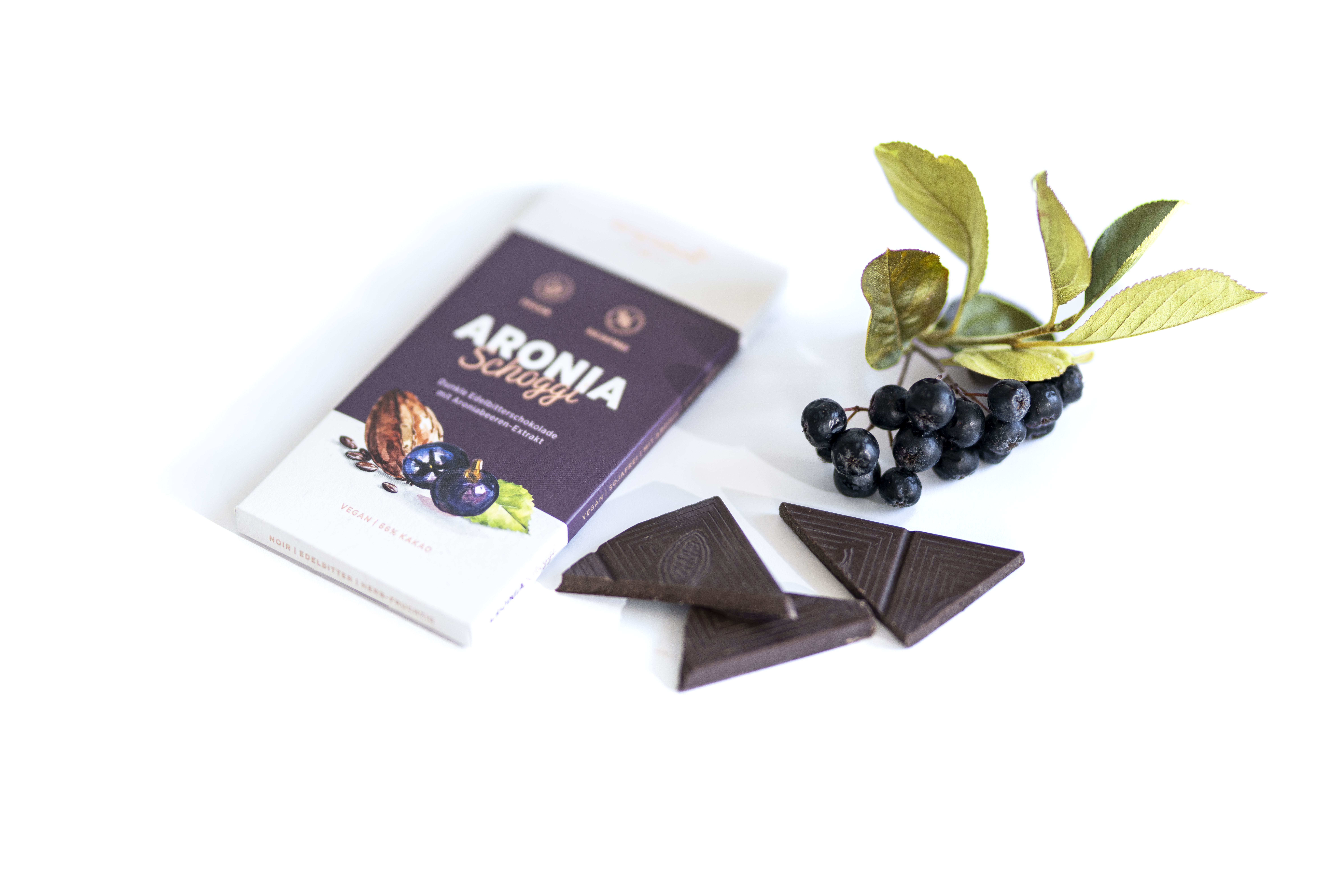 Eine rechteckige Schokoladentafel mit der Aufschrift Aronia Schoggi liegt neben drei dreieckigen Stücken dunkler Schokolade und einem kleinen Zweig Aroniabeeren mit grünen Blättern auf weißem Hintergrund.