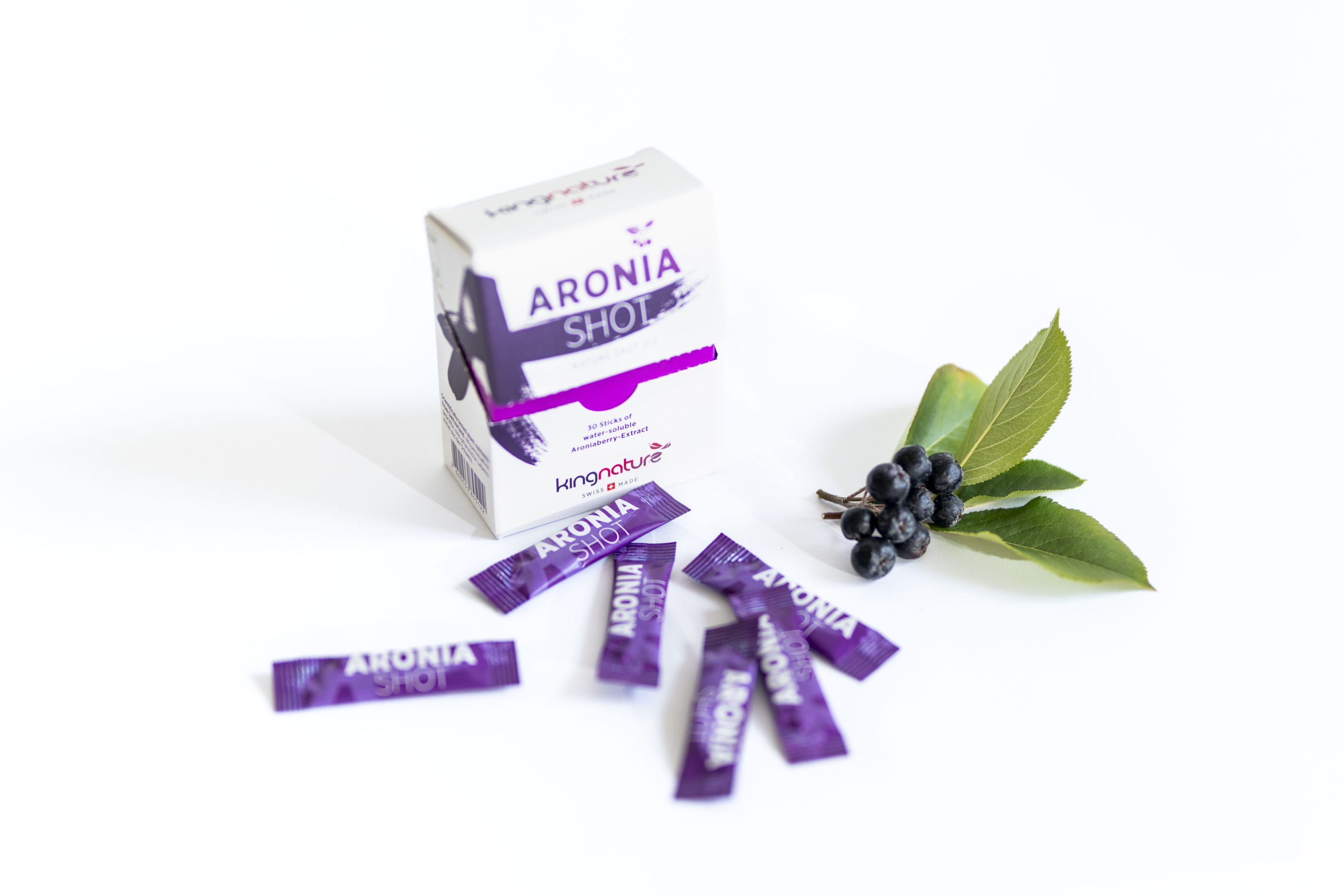 Eine Schachtel mit der Aufschrift "Aronia Shot" mit mehreren violetten Stöcken ist davor ausgebreitet, daneben eine kleine Traube von Aroniabeeren mit grünen Blättern, alles auf weißem Hintergrund.