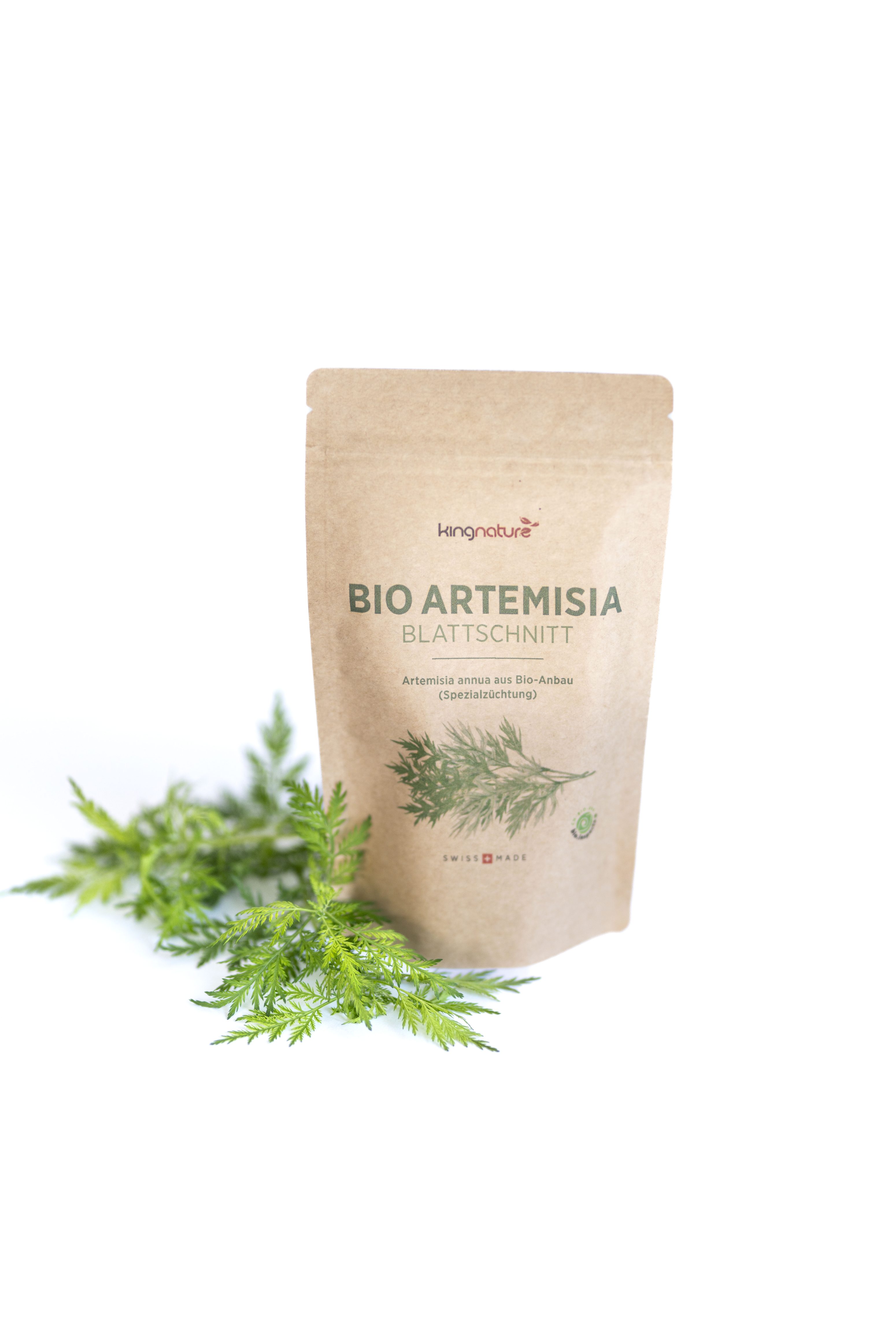 Ein braunes Papierpaket mit der Aufschrift Bio Artemisia Blattschnitt steht aufrecht neben frischen grünen Artemisia-Blättern auf weißem Hintergrund.