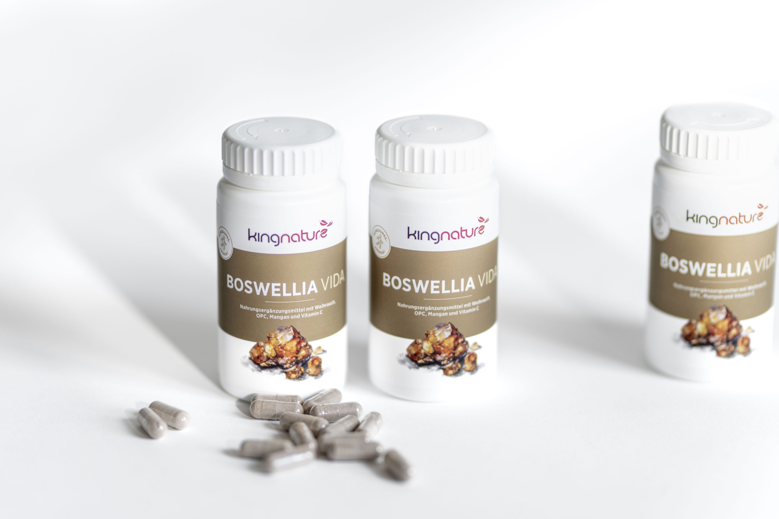 Es werden drei Flaschen mit der Aufschrift kingnature BOSWELLIA VIDA gezeigt, vor denen einige graue Kapseln auf einer weißen Fläche verstreut sind, was auf ein pflanzliches Ergänzungsprodukt schließen lässt.
