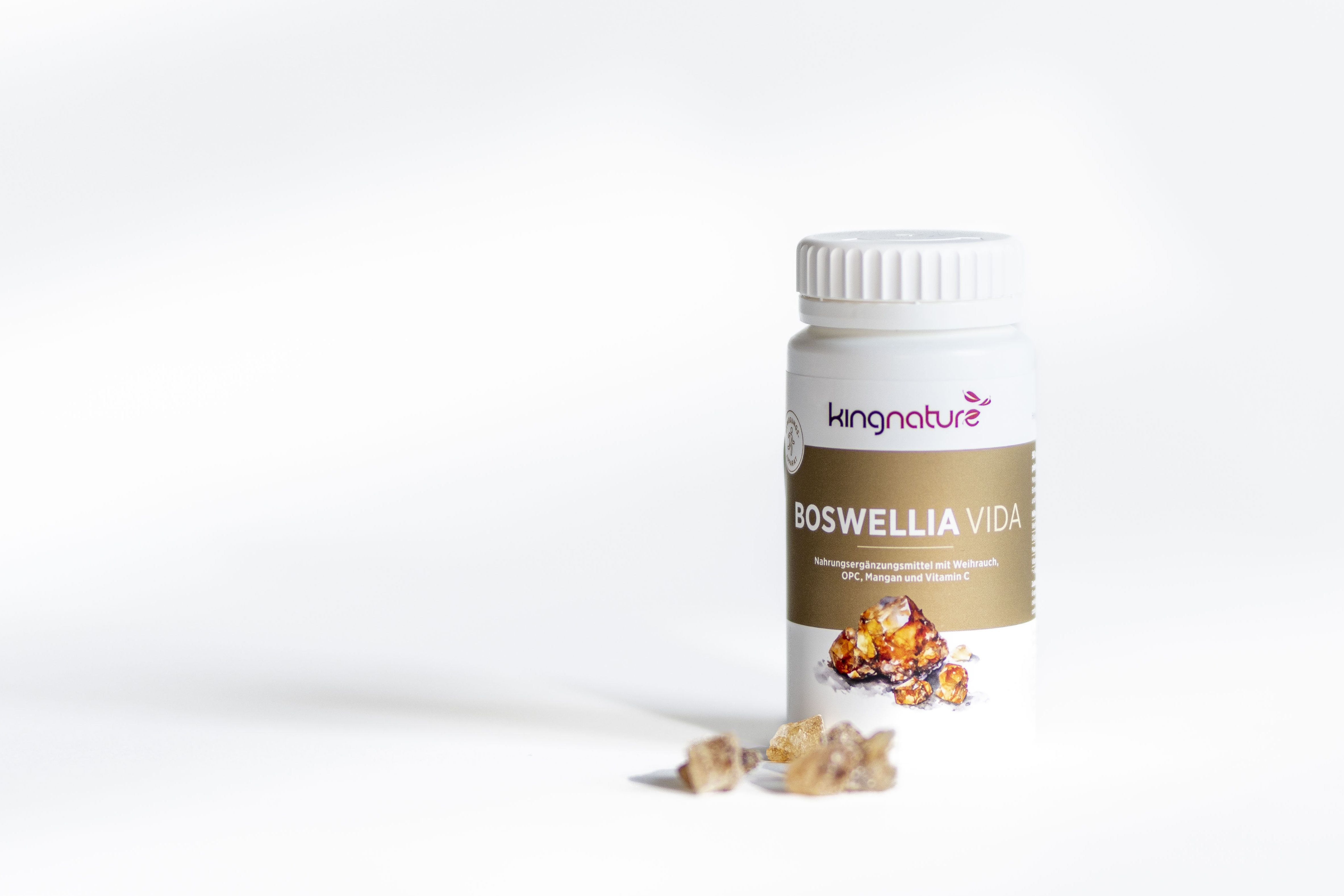 Ein weißes Fläschchen mit der Aufschrift BOSWELLIA VIDA steht auf einer weißen Fläche, neben der einige Stücke getrockneten Boswelliaharzes verstreut sind. Der Hintergrund ist hell und minimalistisch.