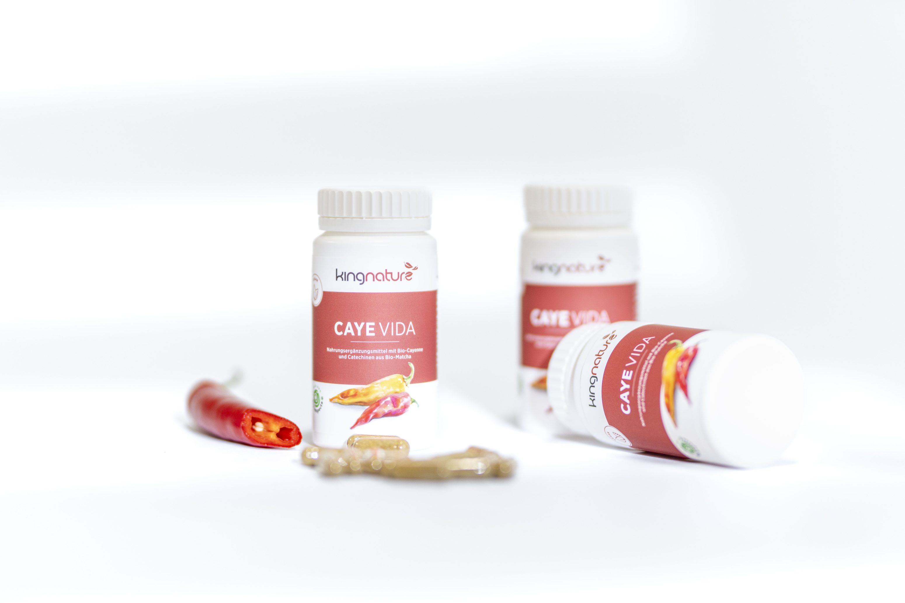 Drei Flaschen von Kingnature Caye Vida Nahrungsergänzungsmitteln sind mit einer roten Chilischote und mehreren Kapseln auf einer weißen Fläche abgebildet, alles hell beleuchtet vor einem schlichten weißen Hintergrund.