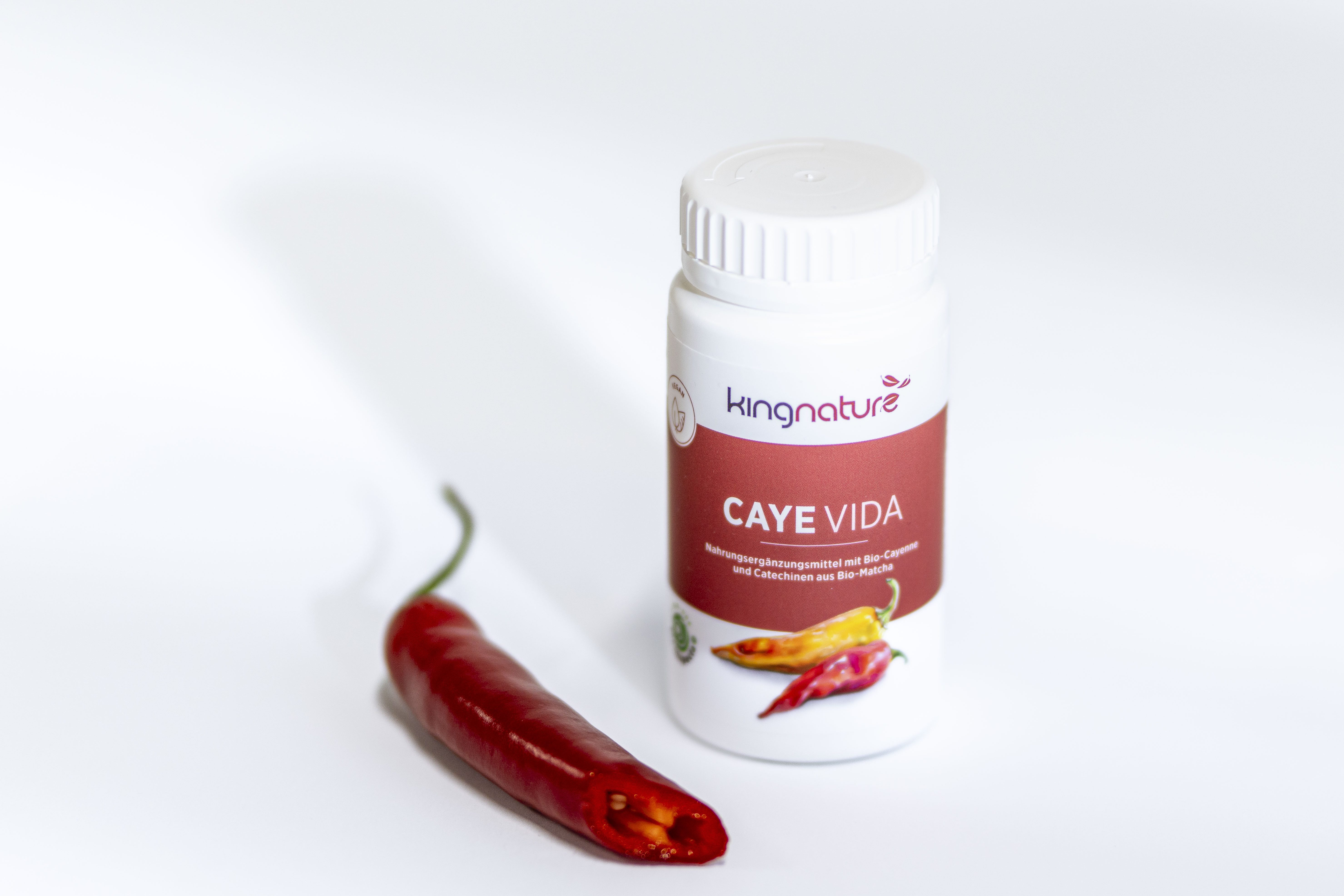Eine weiße Flasche mit der Aufschrift Cayé Vida von Kingnature steht auf einer weißen Fläche neben einer roten Chilischote. Das Etikett zeigt Bilder von Chilischoten und ein rotes Farbschema.