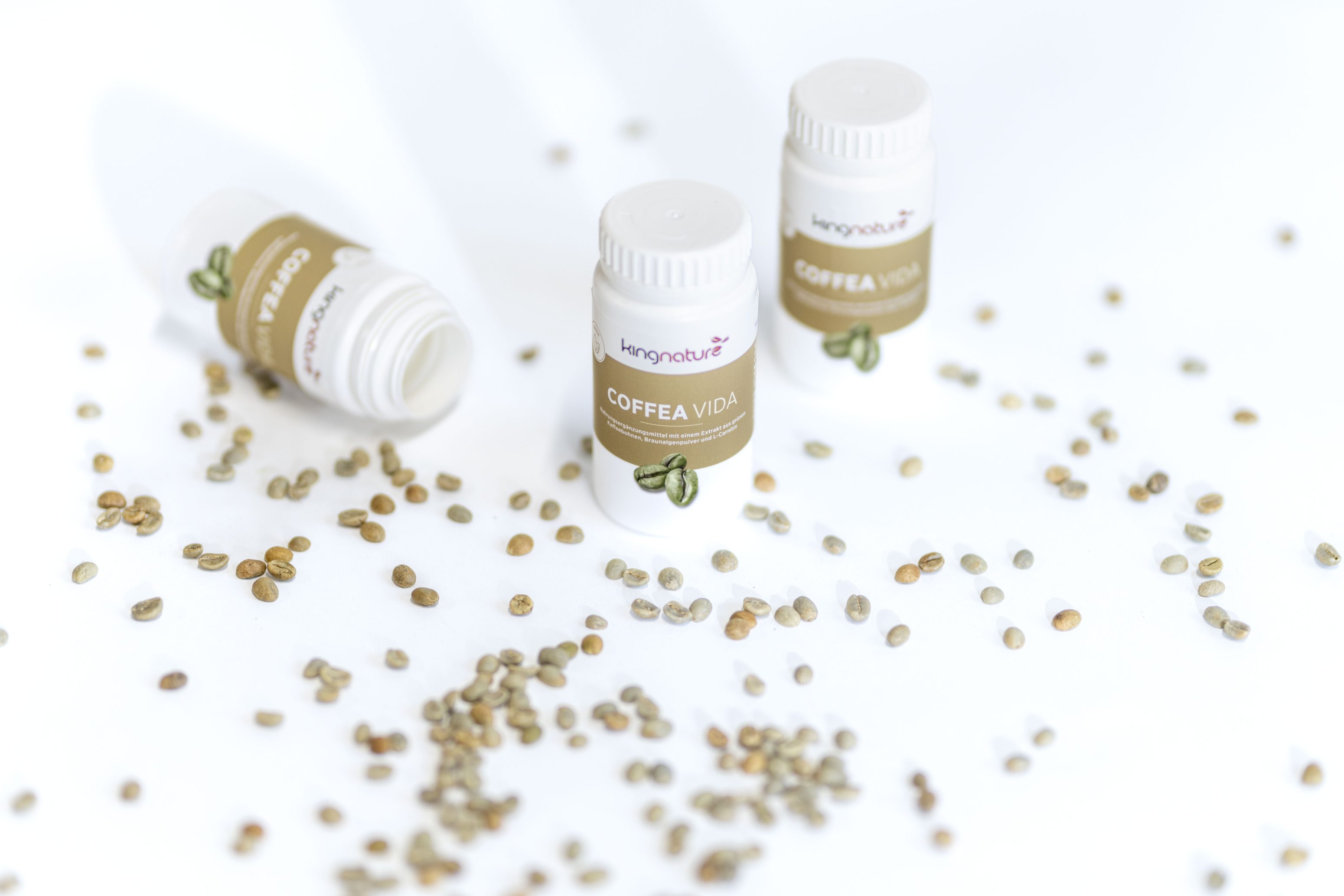 Drei weiße Flaschen mit der Aufschrift Coffea Vida sind von verstreuten Kaffeebohnen auf einer weißen Fläche umgeben. Eine Flasche liegt, während die beiden anderen aufrecht stehen. Auf den Etiketten ist eine grüne Kaffeebohne abgebildet.