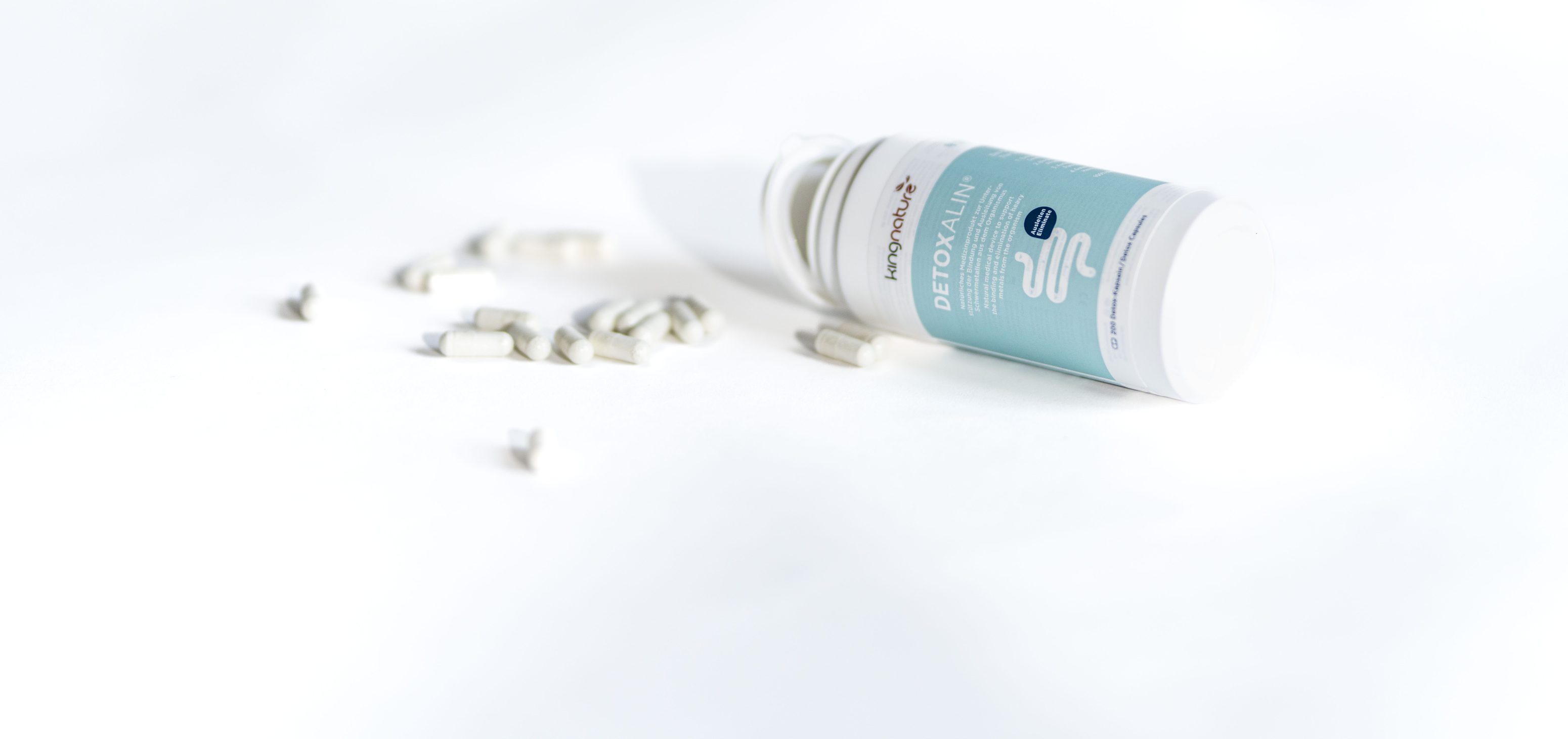 Ein weißes Tablettenfläschchen mit der Aufschrift Detoxalin liegt auf einer weißen Fläche auf der Seite, und in der Nähe sind mehrere weiße Tabletten verstreut. Der Hintergrund ist minimal und hell.