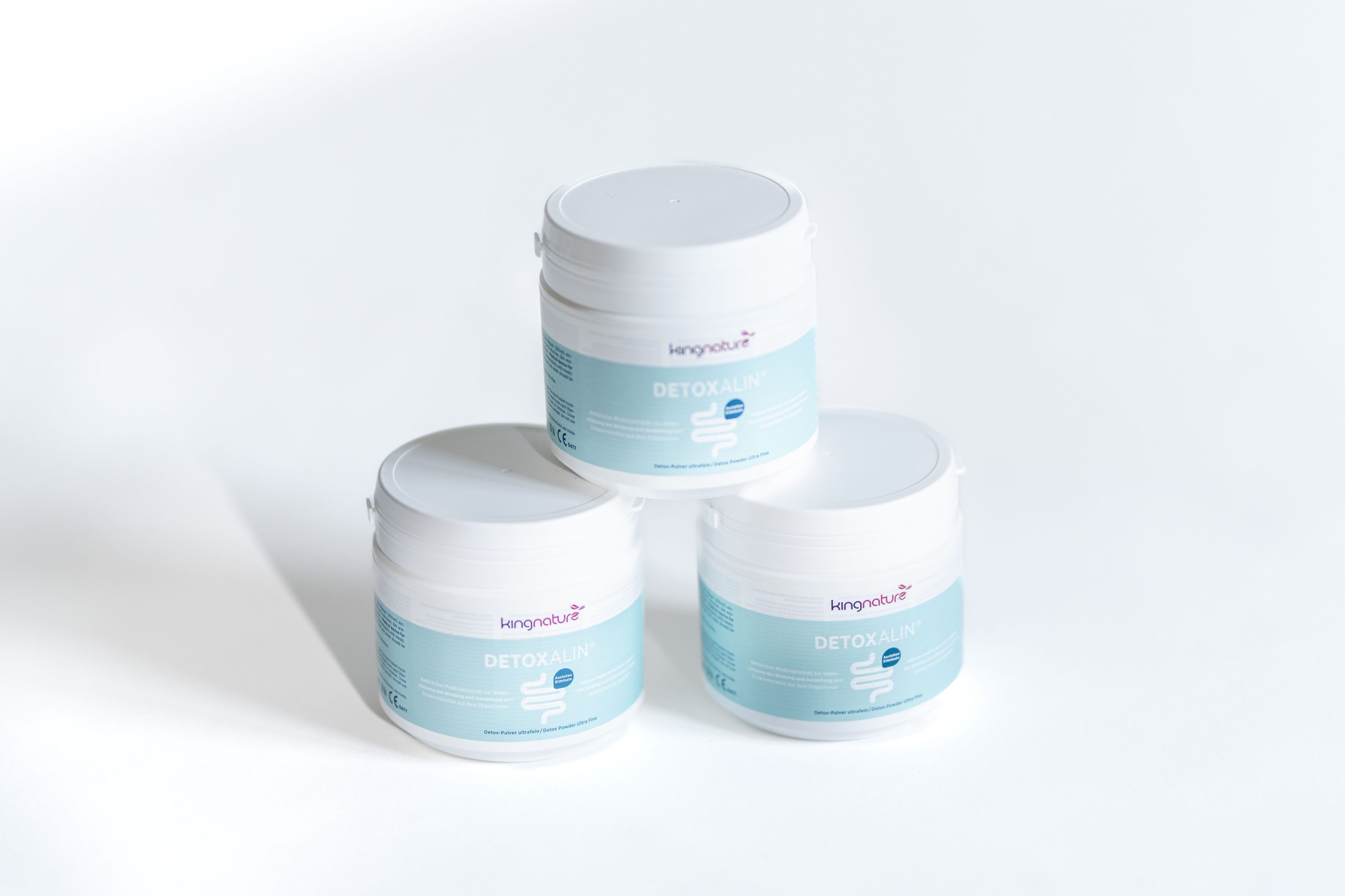 Drei weiße Kunststoffbehälter mit dem Nahrungsergänzungsmittel Detoxalin der Marke Kengouth, die pyramidenförmig auf einer weißen Fläche angeordnet sind, wobei auf jedem Behälter ein hellblaues Etikett sichtbar ist.