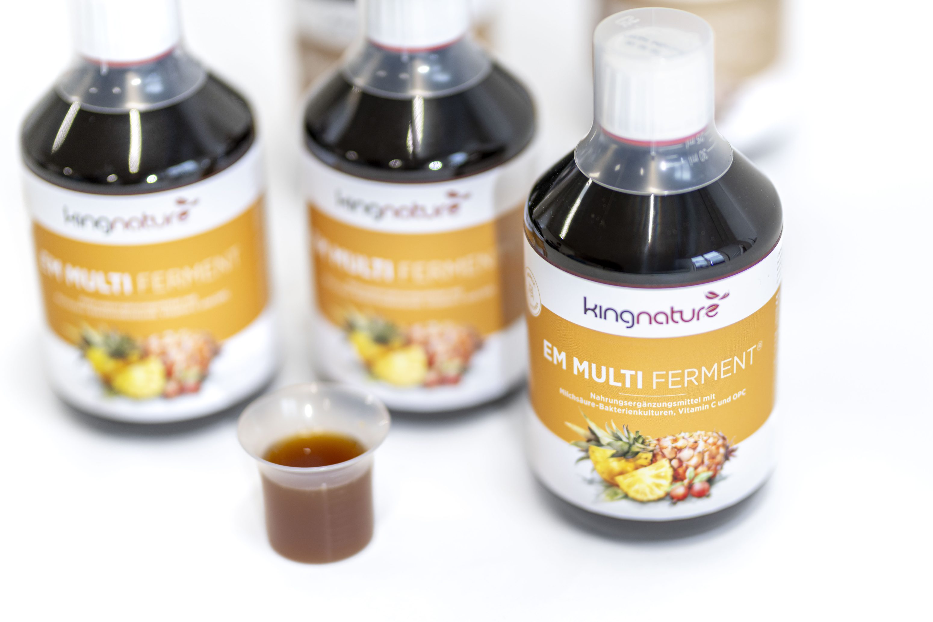 Drei Flaschen des Nahrungsergänzungsmittels Kingnature EM Multi Ferment mit weißen Verschlüssen und bunten Etiketten mit Ananas- und Granatapfelmotiven stehen auf einer weißen Fläche; davor steht ein kleiner Becher mit der Flüssigkeit.