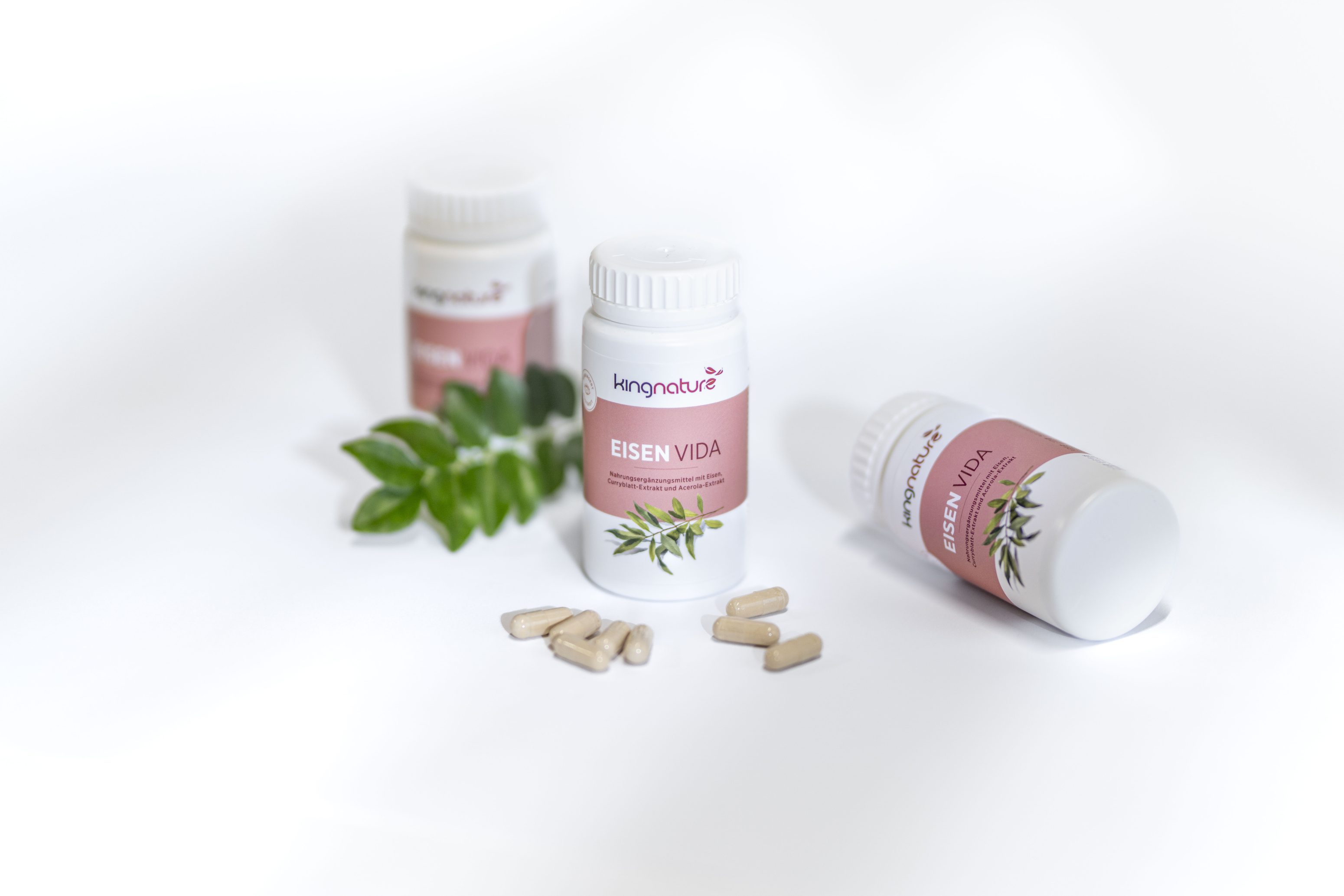 Drei weiße Flaschen mit der Aufschrift Eisen Vida mit rosa Etiketten und grünen Blättern sind auf einer weißen Fläche ausgestellt, daneben mehrere beigefarbene Kapseln mit grünen Blättern. Der Markenname kingnature ist auf jeder Flasche sichtbar.