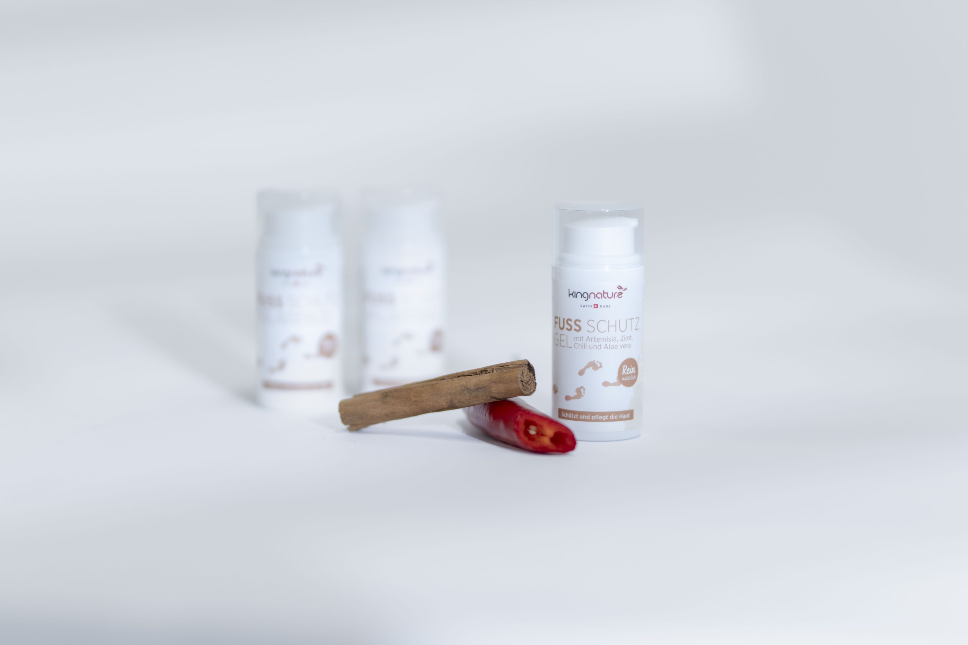 Eine Flasche mit RUSS SCHUTZ-Kapseln steht im Mittelpunkt, davor eine Zimtstange und eine rote Chilischote. Zwei ähnliche Flaschen stehen unscharf im Hintergrund auf einer weißen Fläche.