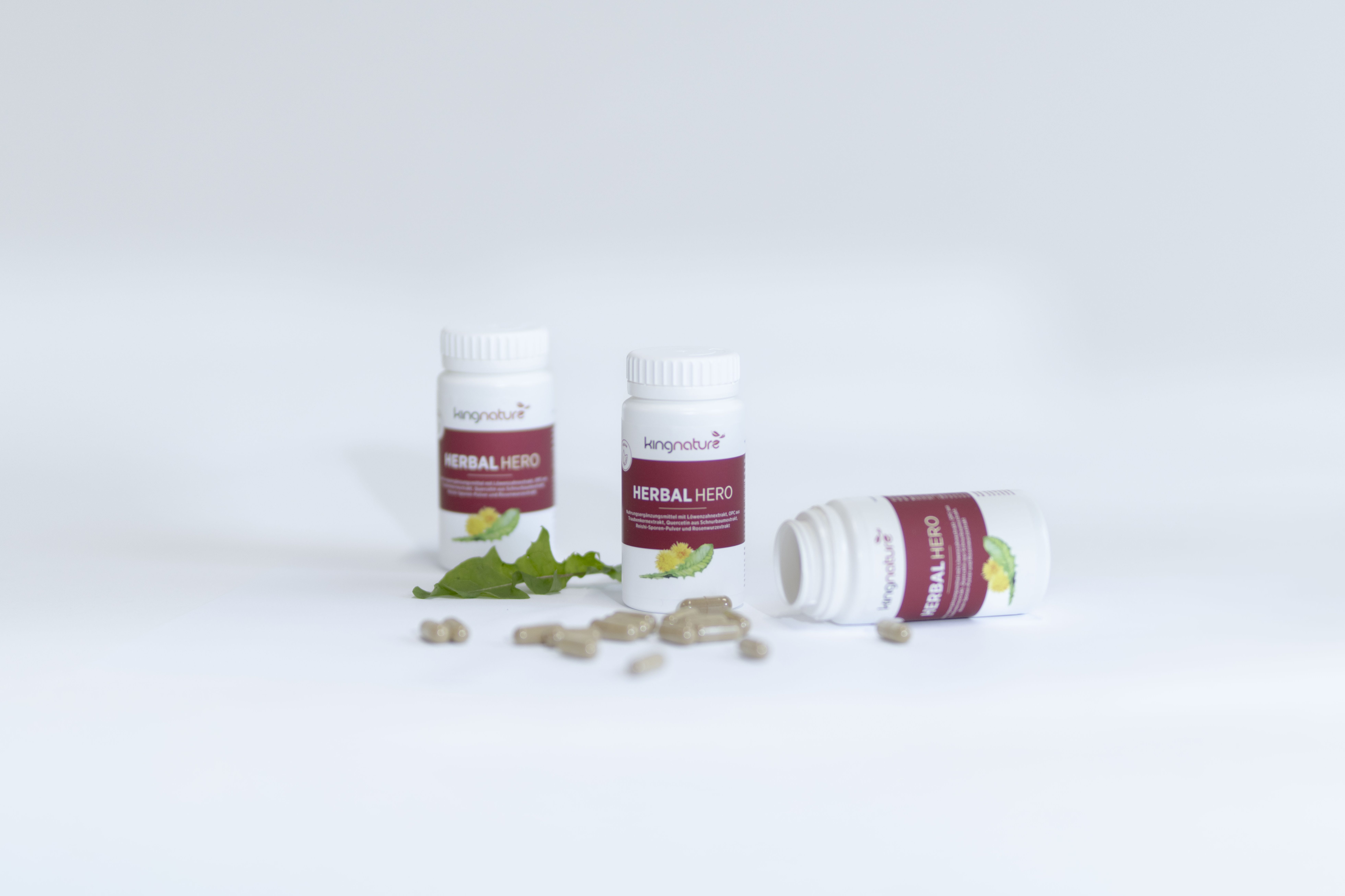 Drei Flaschen mit Kräuterkapseln mit der Aufschrift Herbal Hero sind abgebildet, wobei eine Flasche auf der Seite liegt und zwei aufrecht stehen. Die Kapseln und grünen Blätter sind auf einer weißen Fläche verstreut.
