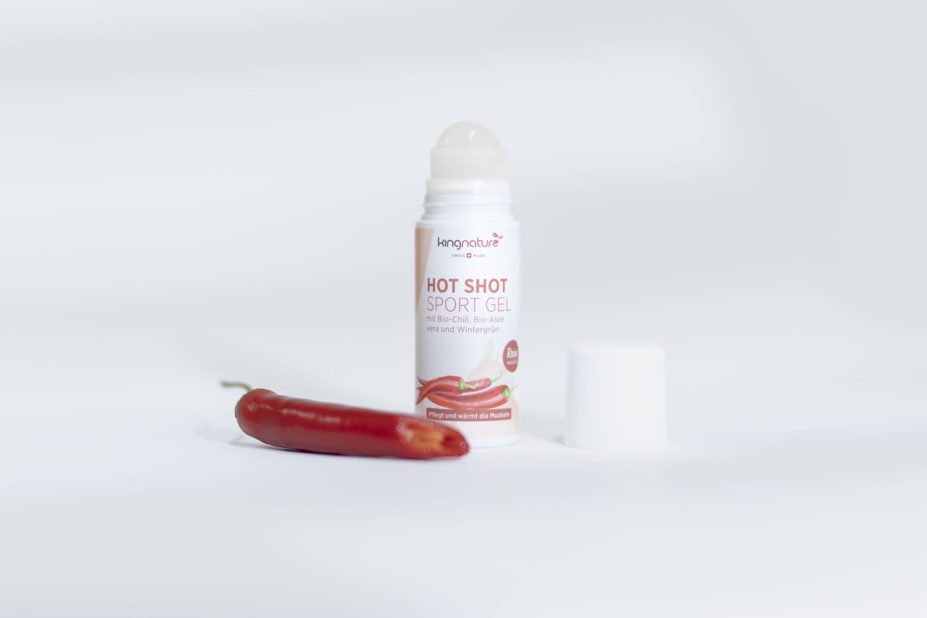 Ein Roll-on-Fläschchen von Kingnature Hot Shot Sport Gel mit abgenommener Kappe steht aufrecht neben einer roten Chilischote auf einer weißen Fläche mit minimalistischem Hintergrund.