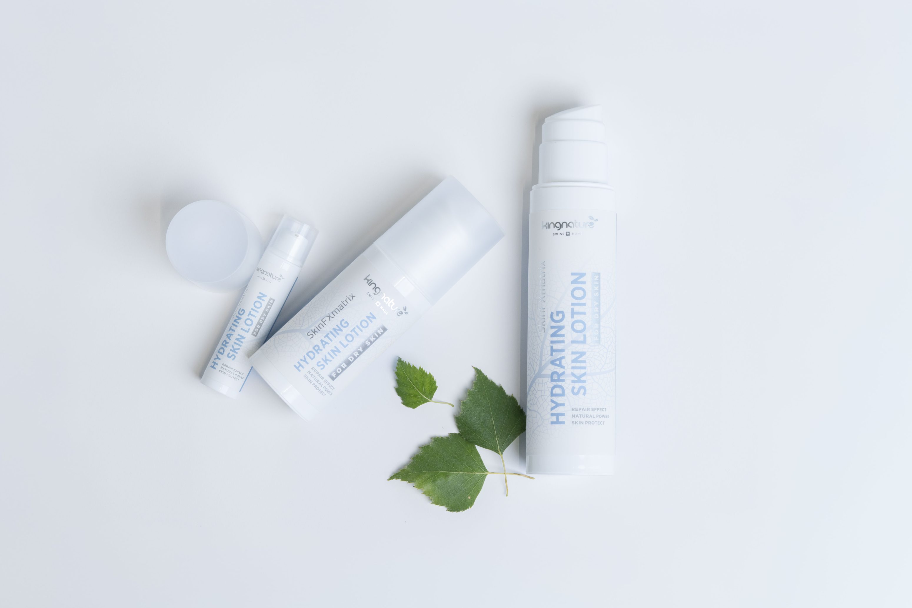 Drei weiße Hautpflegeproduktflaschen mit der Aufschrift Hydrating Skin Lotion sind auf einer weißen Fläche mit zwei grünen Blättern daneben angeordnet, wodurch eine saubere und frische Präsentation entsteht.