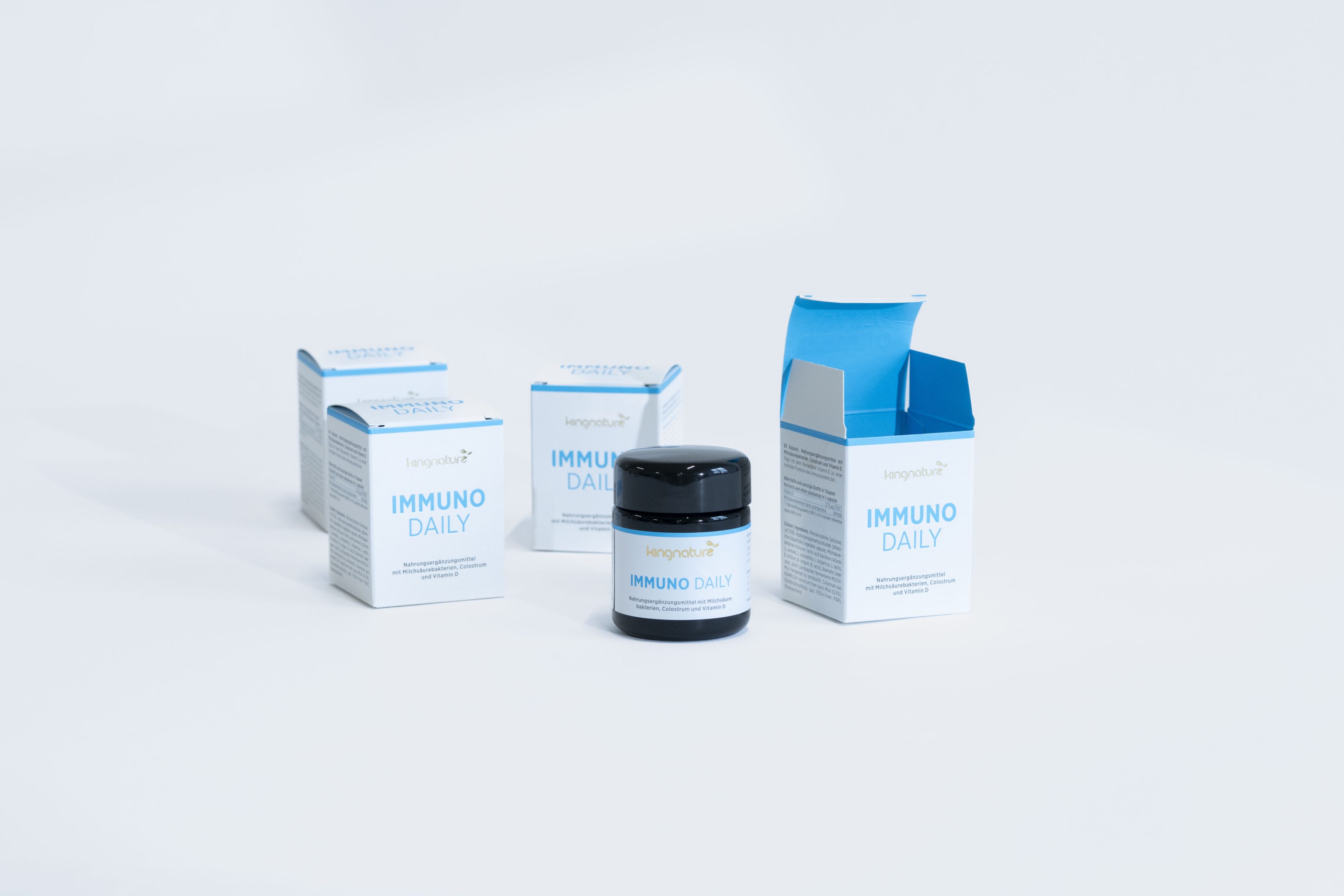 Vier Schachteln und eine Dose des Nahrungsergänzungsmittels Immuno Daily mit weißer und blauer Verpackung sind auf weißem Hintergrund angeordnet. Eine Schachtel ist geöffnet und gibt den Blick auf die Dose frei.