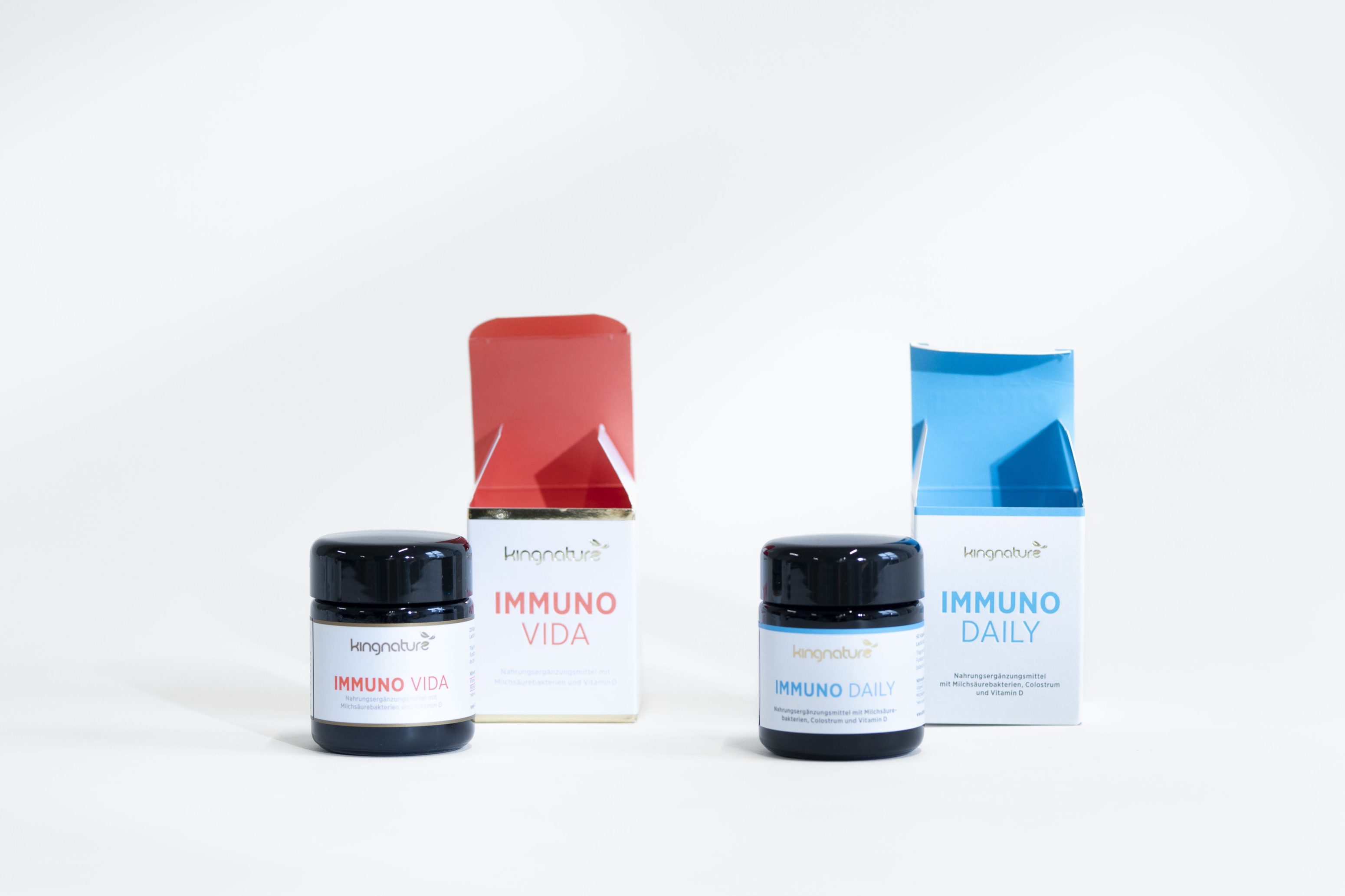 Auf weißem Hintergrund sind zwei Nahrungsergänzungsdosen mit den dazugehörigen Schachteln abgebildet. Das linke Glas und die Schachtel sind mit IMMUNO VIDA in Rot beschriftet, das rechte Set mit IMMUNO DAILY in Blau.