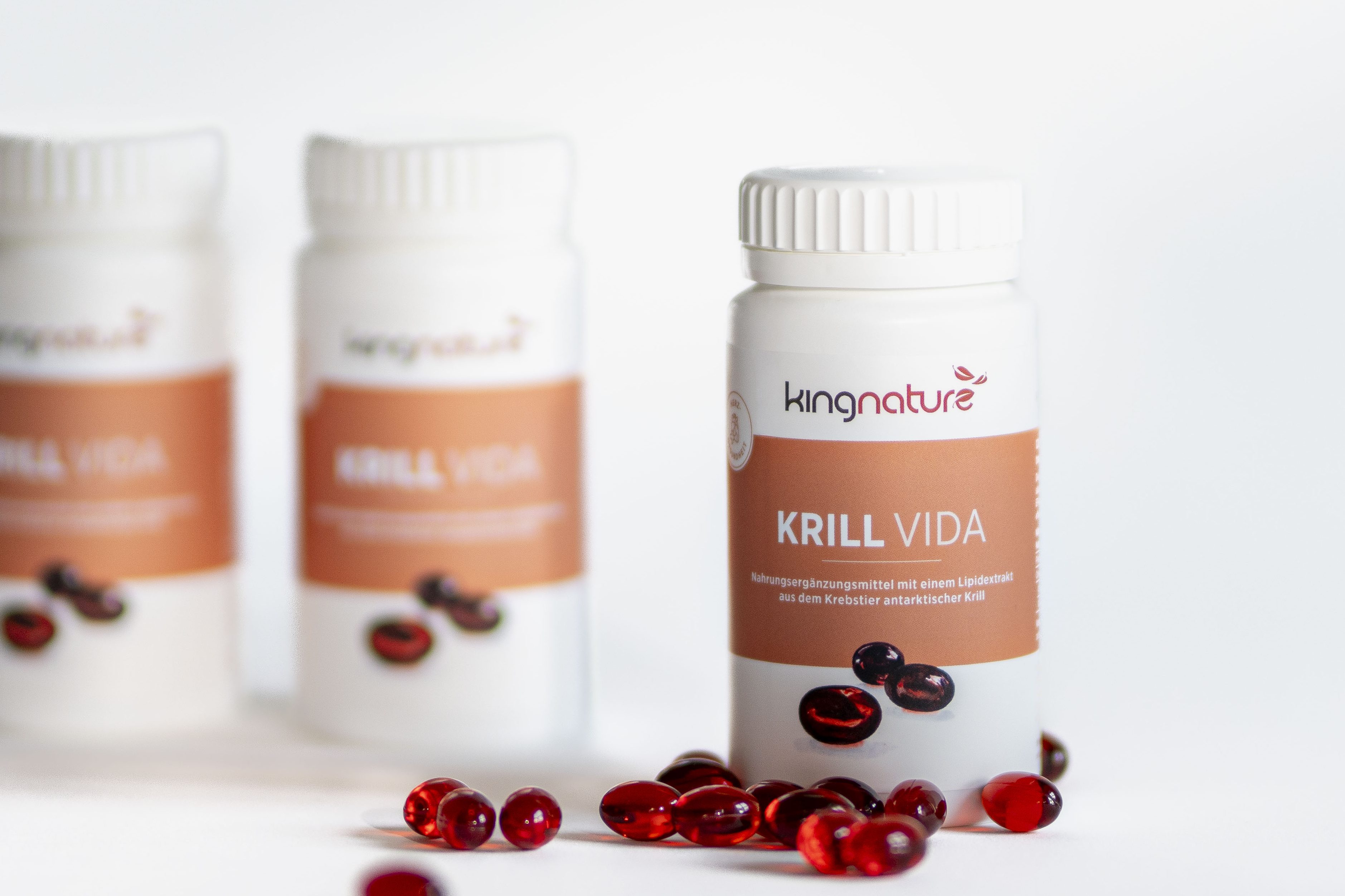 Drei weiße Flaschen mit der Aufschrift Krill Vida stehen auf einer weißen Fläche, umgeben von verstreuten roten Kapseln. Zwei Flaschen sind im Hintergrund unscharf, während eine im Vordergrund deutlich zu sehen ist.