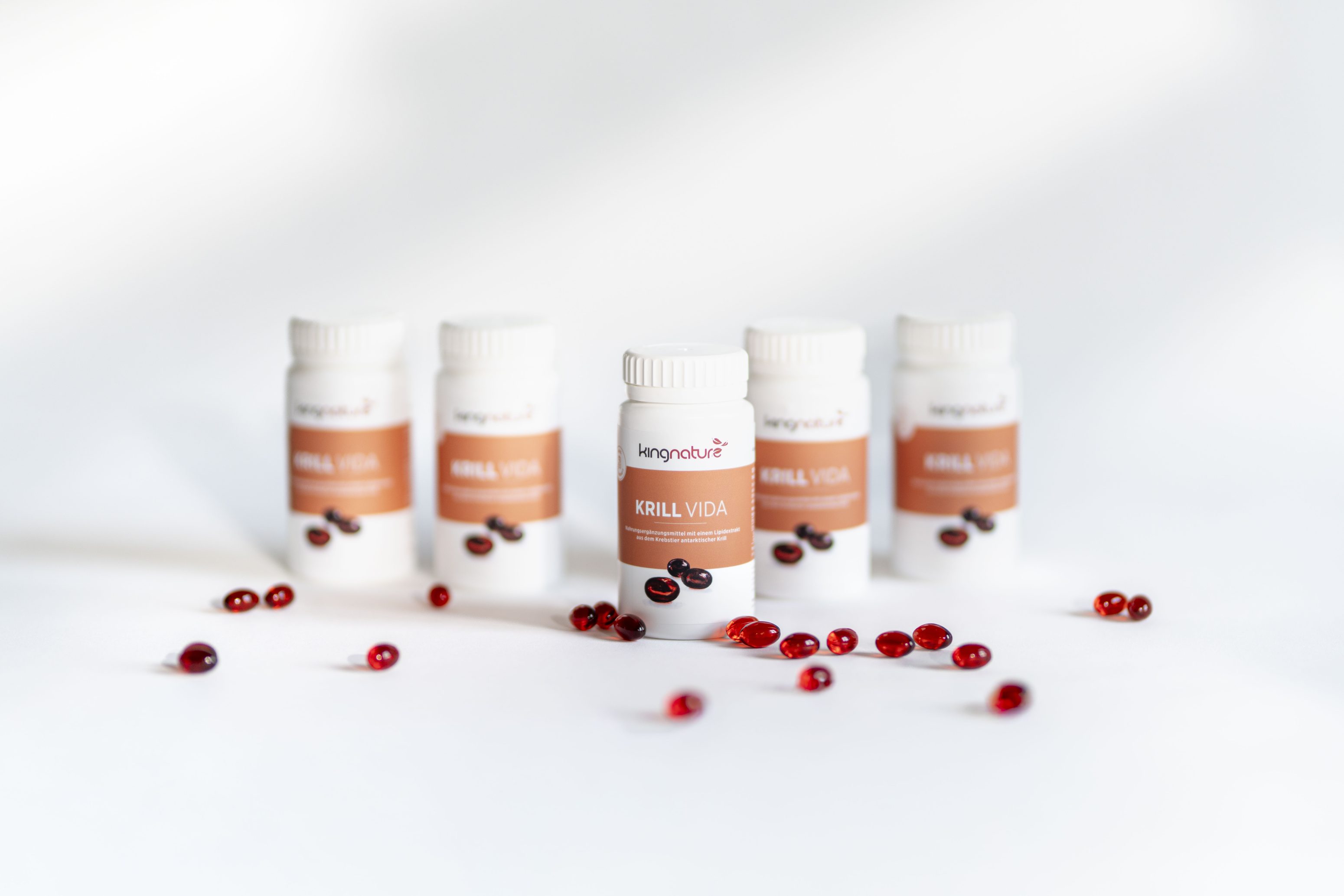 Fünf Flaschen mit der Aufschrift "KRILL VIDA", um die herum rote Kapseln verstreut sind; nur die Flasche in der Mitte ist scharf eingestellt, während die anderen auf einer weißen Fläche im Hintergrund verschwimmen.