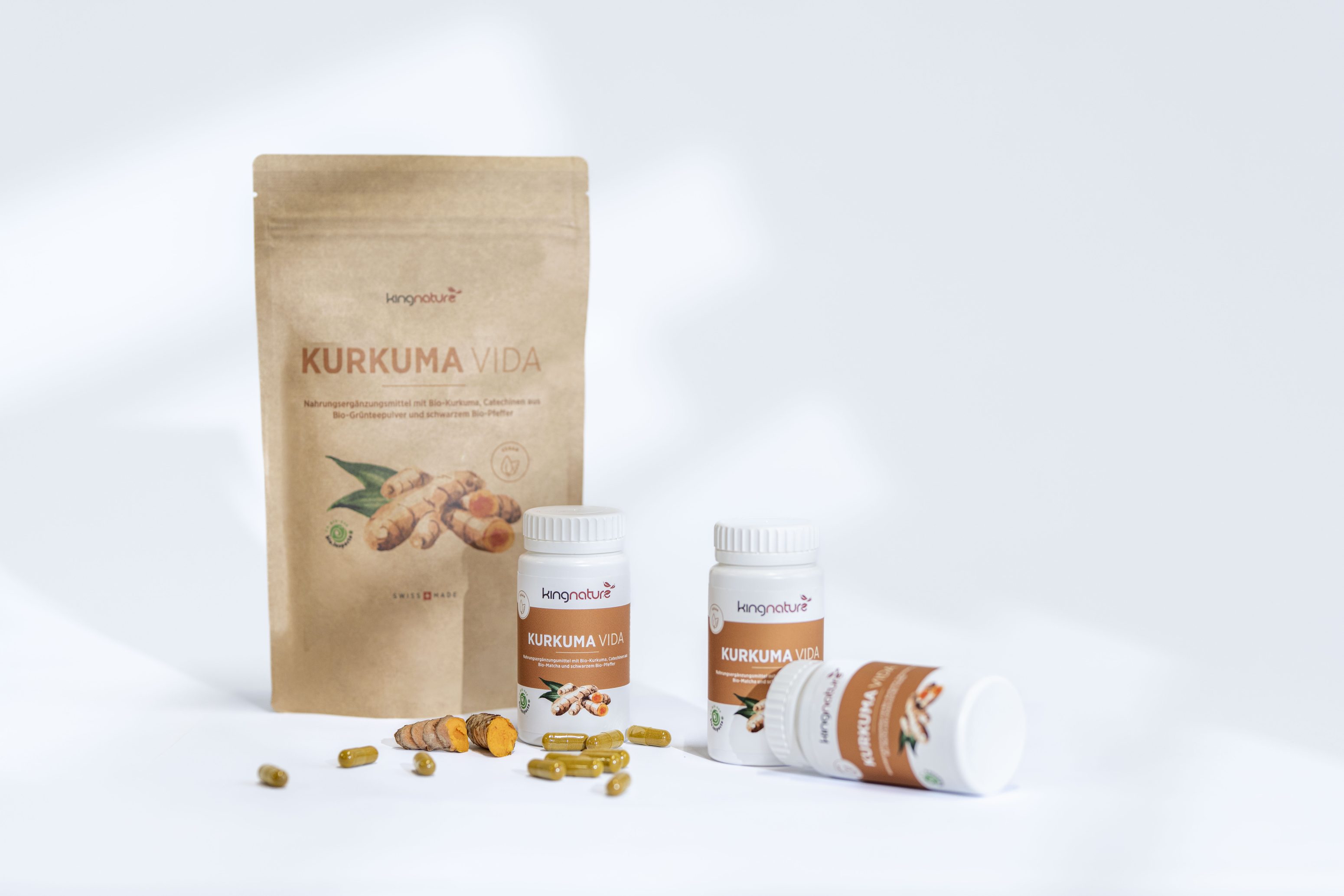 Ein Beutel und zwei Flaschen mit der Aufschrift "Kurkuma Vida" und Abbildungen von Kurkumawurzeln sind neben mehreren geöffneten gelben Kapseln und einigen losen Kurkumakapseln auf weißem Hintergrund zu sehen.