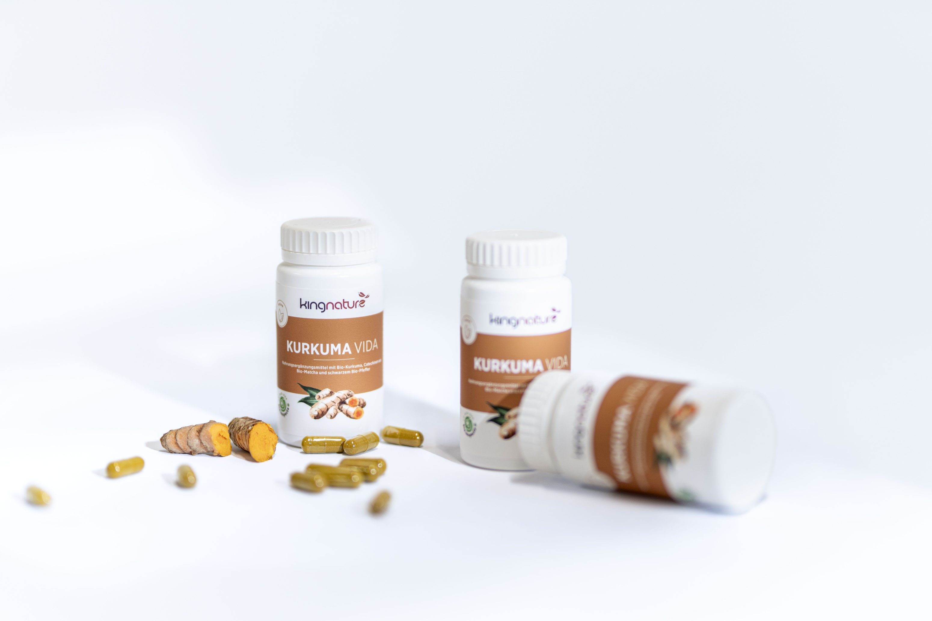 Drei weiße Flaschen mit der Aufschrift Kurkuma Vida, um die herum Kurkumakapseln und Kurkumawurzelscheiben auf weißem Hintergrund verstreut sind. Eine Flasche liegt auf der Seite.