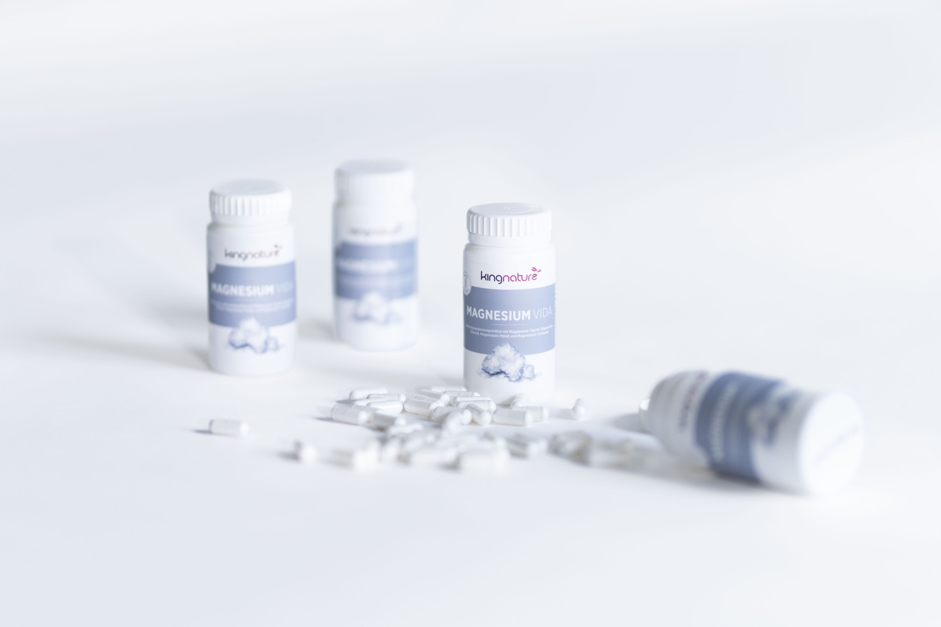 Vier weiße Flaschen mit der Aufschrift Magnesium stehen auf einer weißen Fläche, wobei eine Flasche auf der Seite liegt und mehrere weiße Tabletten vor ihr verteilt sind. Der Hintergrund ist sanft verschwommen.