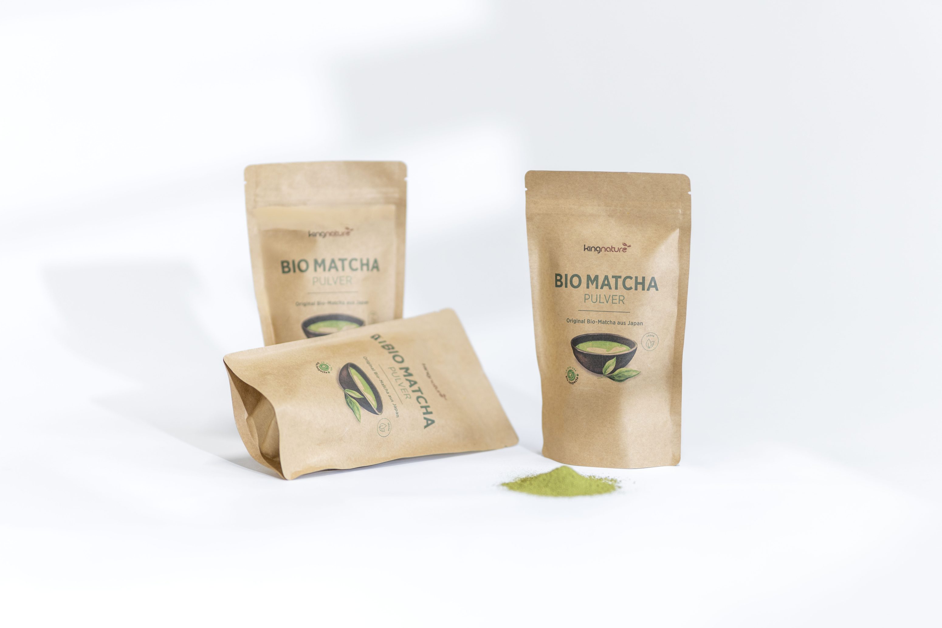 Drei braune Papierbeutel mit der Aufschrift "Bio Matcha Pulver" werden mit grünem Matcha-Pulver bestreut präsentiert. Zwei Beutel stehen aufrecht, und ein Beutel liegt auf der Seite auf weißem Hintergrund.