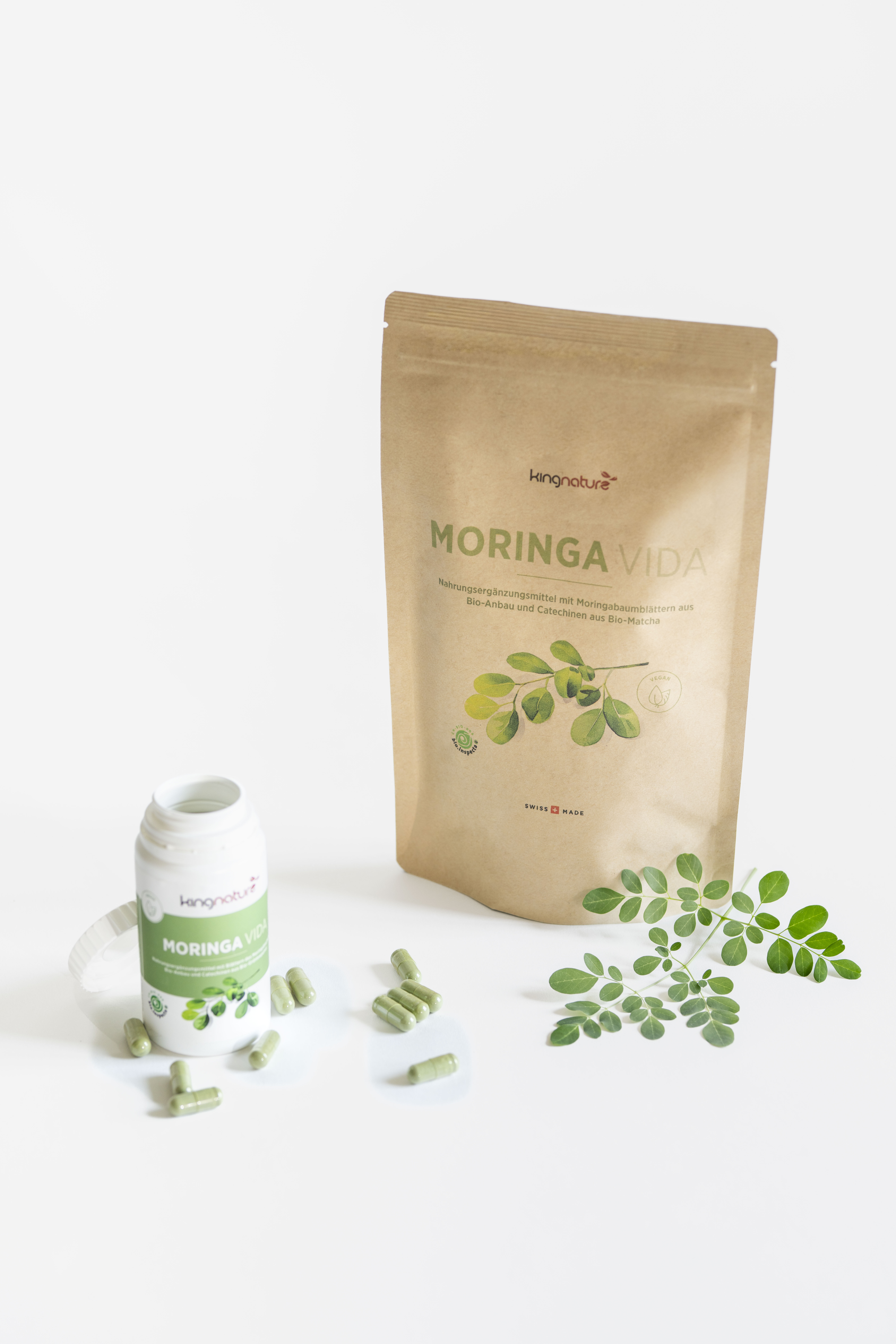 Eine Flasche und ein Beutel mit der Aufschrift "Moringa Vida" sind mit grünen Kapseln, einer weißen Kappe und Moringa-Blättern auf einer weißen Fläche versehen. Auf der Verpackung sind grüne Blätter und Text abgebildet.