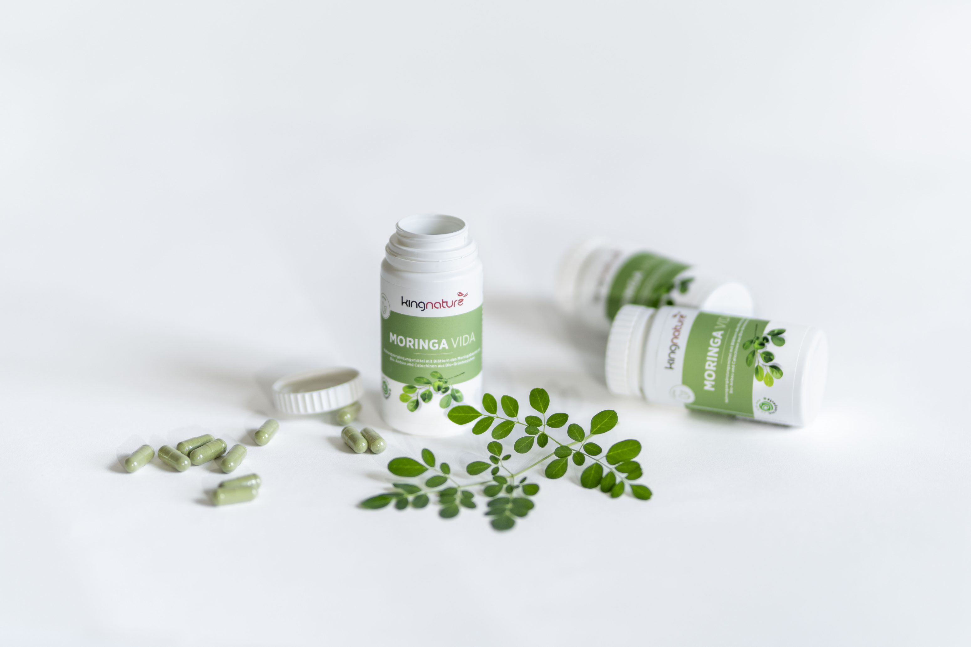 Drei Flaschen mit grünen Etiketten mit der Aufschrift Moringa Vida, von denen eine geöffnet ist und aus der grüne Kapseln herausquellen, stehen auf einer weißen Fläche neben frischen Moringablättern.