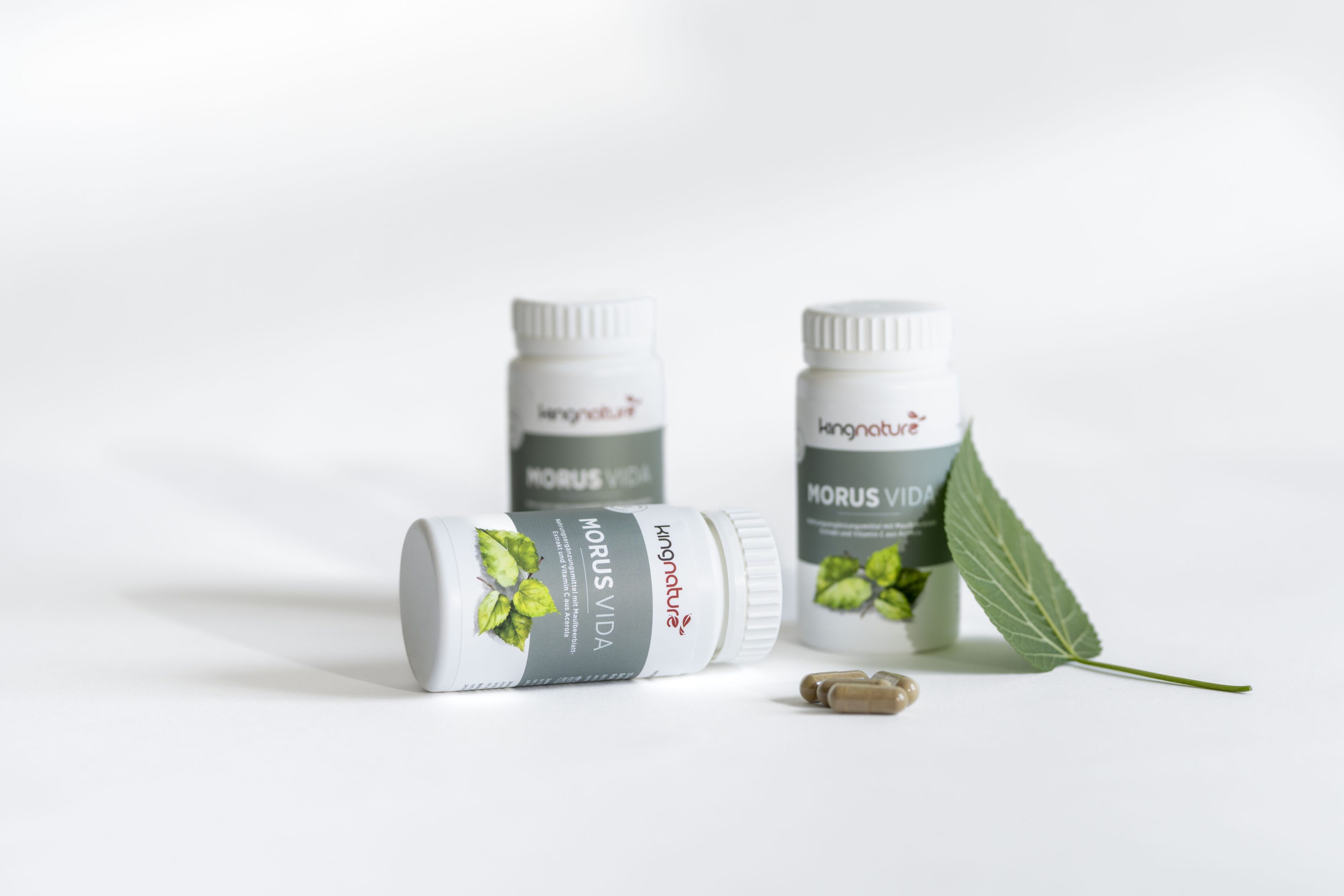 Drei Flaschen mit der Aufschrift Morus Vida von Iamnature stehen auf einer weißen Fläche, wobei eine Flasche auf dem Boden liegt. Daneben befinden sich zwei Kapseln und ein grünes Blatt. Die Szene ist gut beleuchtet und hat einen sauberen Hintergrund.