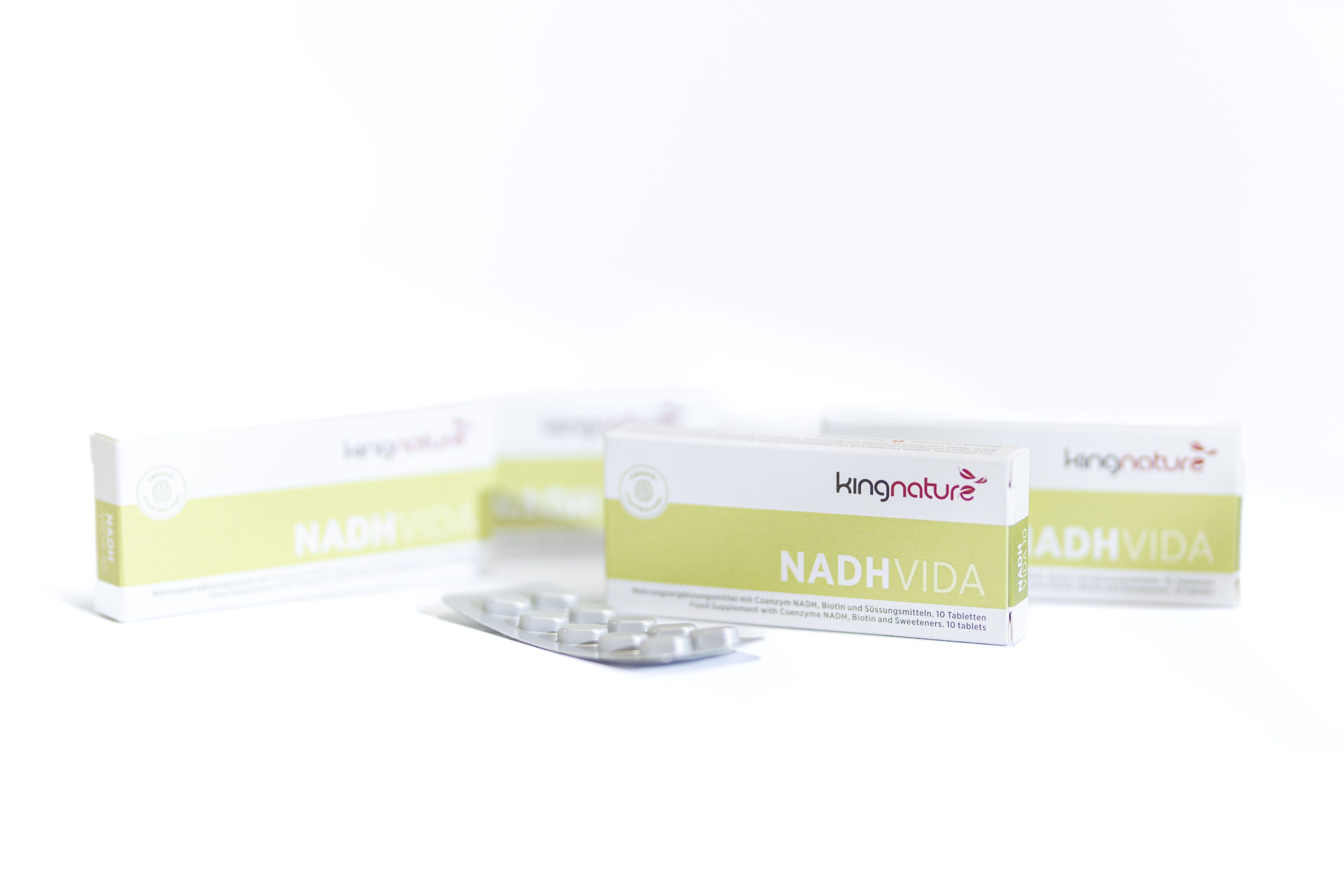 Drei Schachteln mit der Aufschrift NADH VIDA von Kingnature sind auf einer weißen Fläche ausgestellt, wobei eine geöffnete Schachtel eine Blisterpackung mit Tabletten zum Vorschein bringt. Der Hintergrund ist minimal und hell.