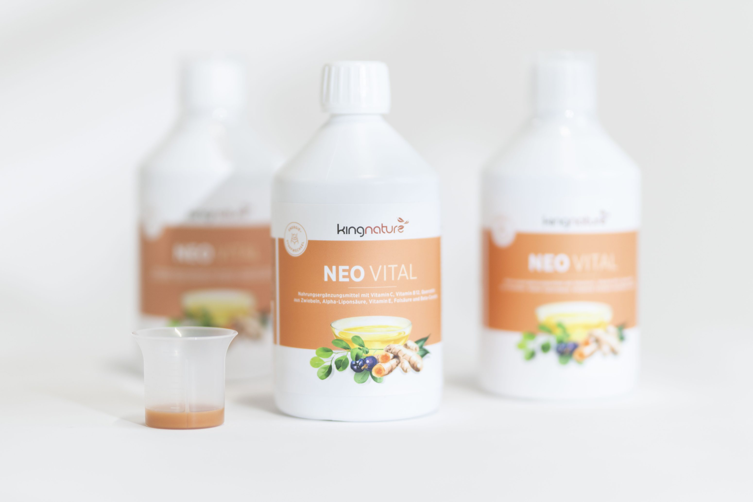 Drei Flaschen des Nahrungsergänzungsmittels Kingnature NEO VITAL mit orangefarbenen und weißen Etiketten sind zu sehen, zwei davon verschwommen im Hintergrund. Davor steht ein kleiner Plastikbecher, der mit einer rötlichen Flüssigkeit gefüllt ist.